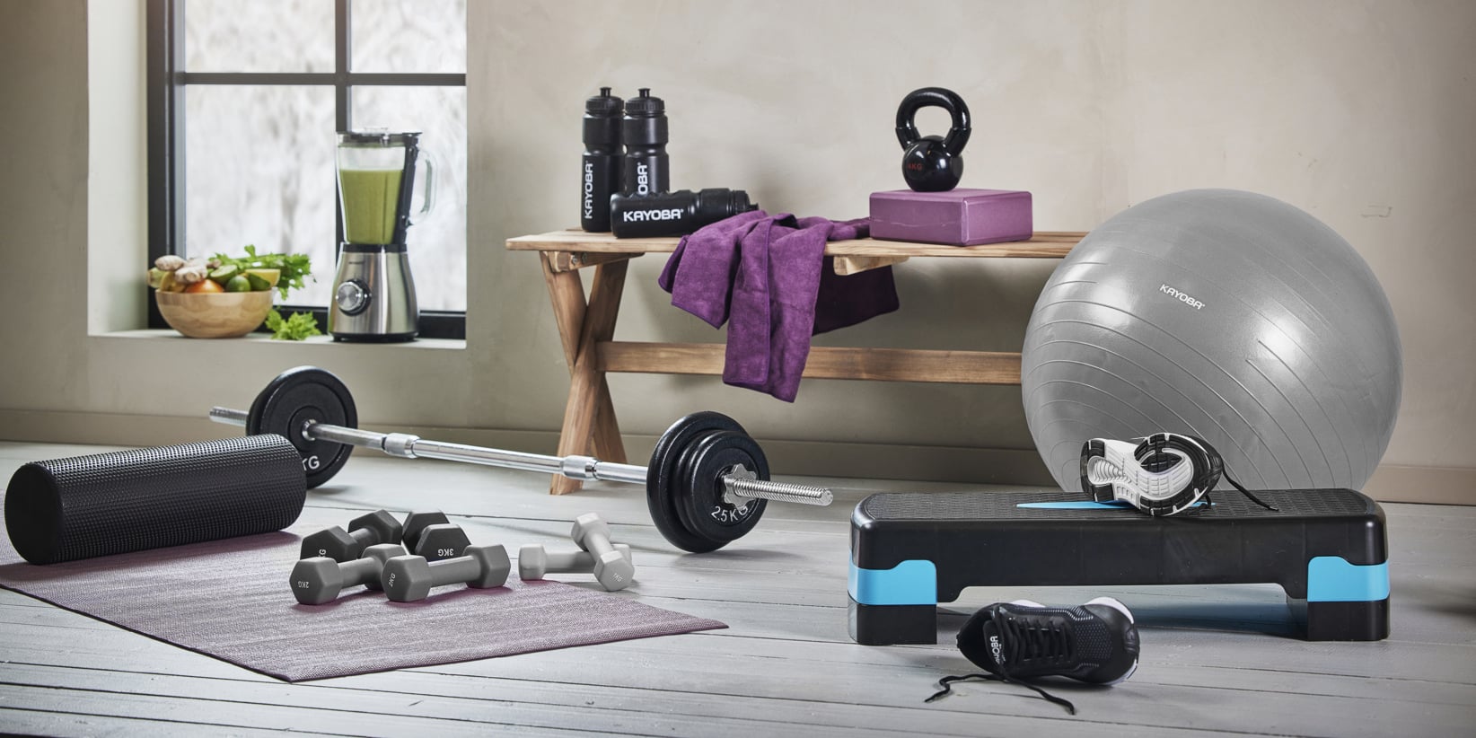 Et hjemmegym ved et vindu har en vektstang, manualer, foam roller, yogamatte, gymball, kettlebell, stepplattform, joggesko, bånd, vannflasker, håndkle, blender og fruktskål.