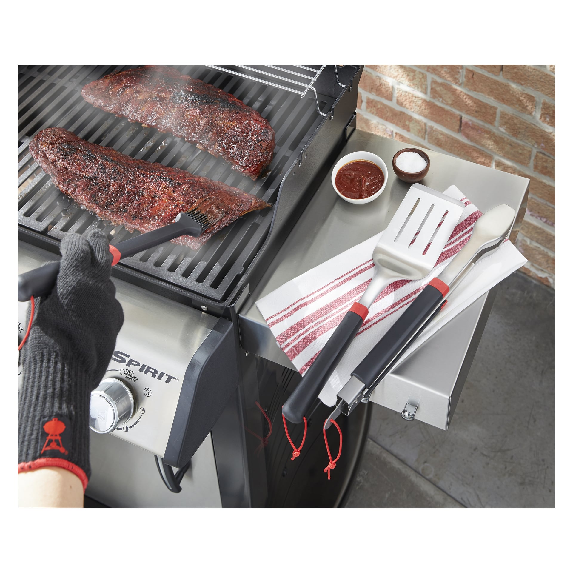 Någon i värmetålig handske borstar sås på revbensgrillning över gas, med bestick, handduk och skålar med sås, salt och peppar i närheten.
