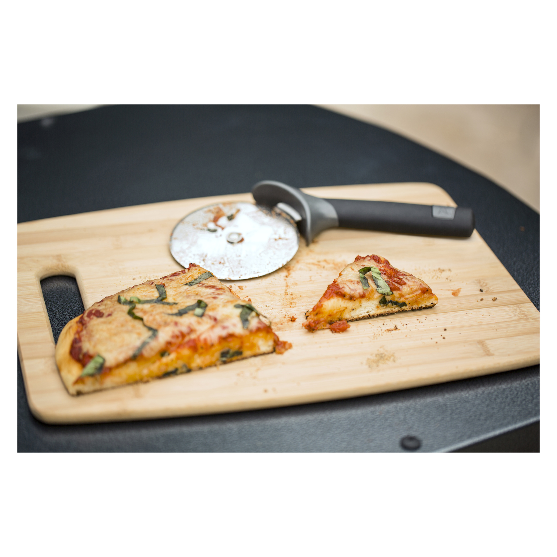 En träskiva håller en delvis skivad pizza med en bit bortdragen och en pizzaskärare bredvid, smulor och örter utspridda omkring.