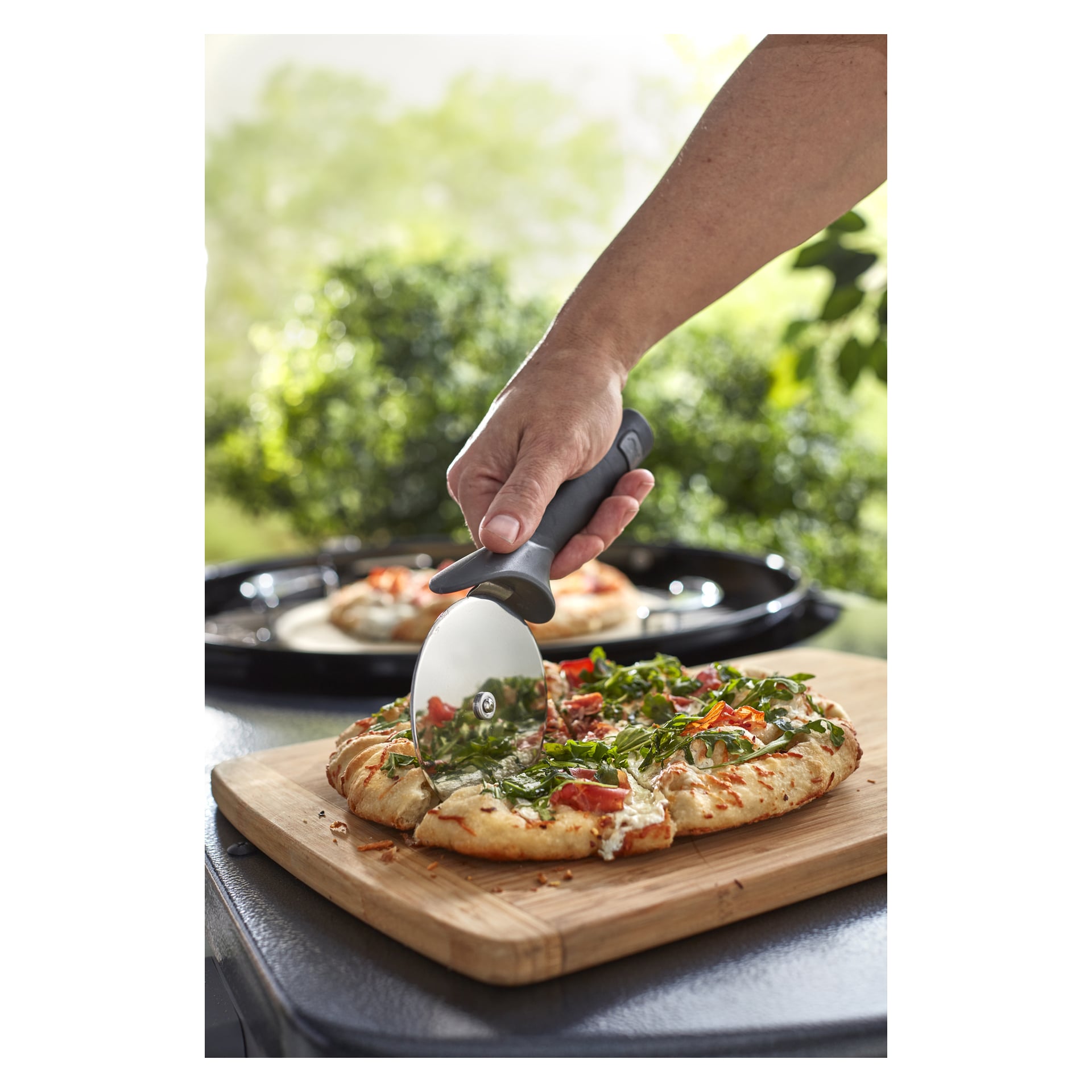 En hand skär en vegetarisk pizza med en skärare på en träskiva, medan en annan pizza ligger bakom på en svart bricka bland utomhusgrönska.