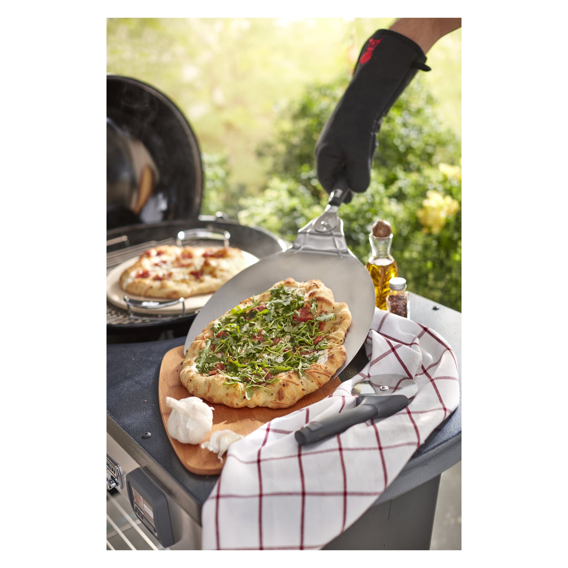 Någon i svart ugnsvante håller i ett pizzaskal med en pizza toppad med grönt topp; En annan bakas på grillen. Handduk, vitlök, olja och peppar i närheten.