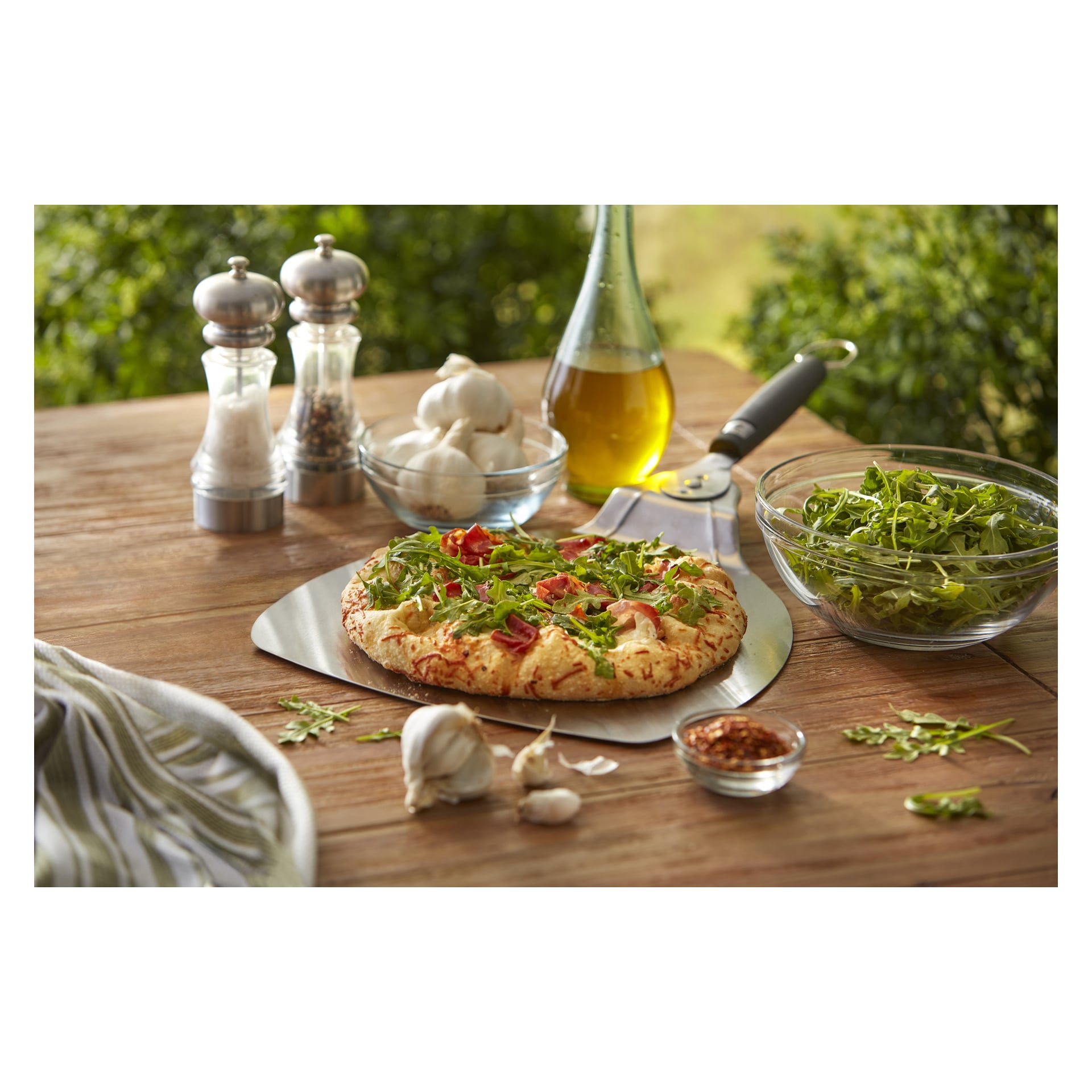 En vegetarisk pizza med ruccola står på ett skal på ett träbord, nära vitlök, olivolja, sallad, shakers och en pizzaskärare.