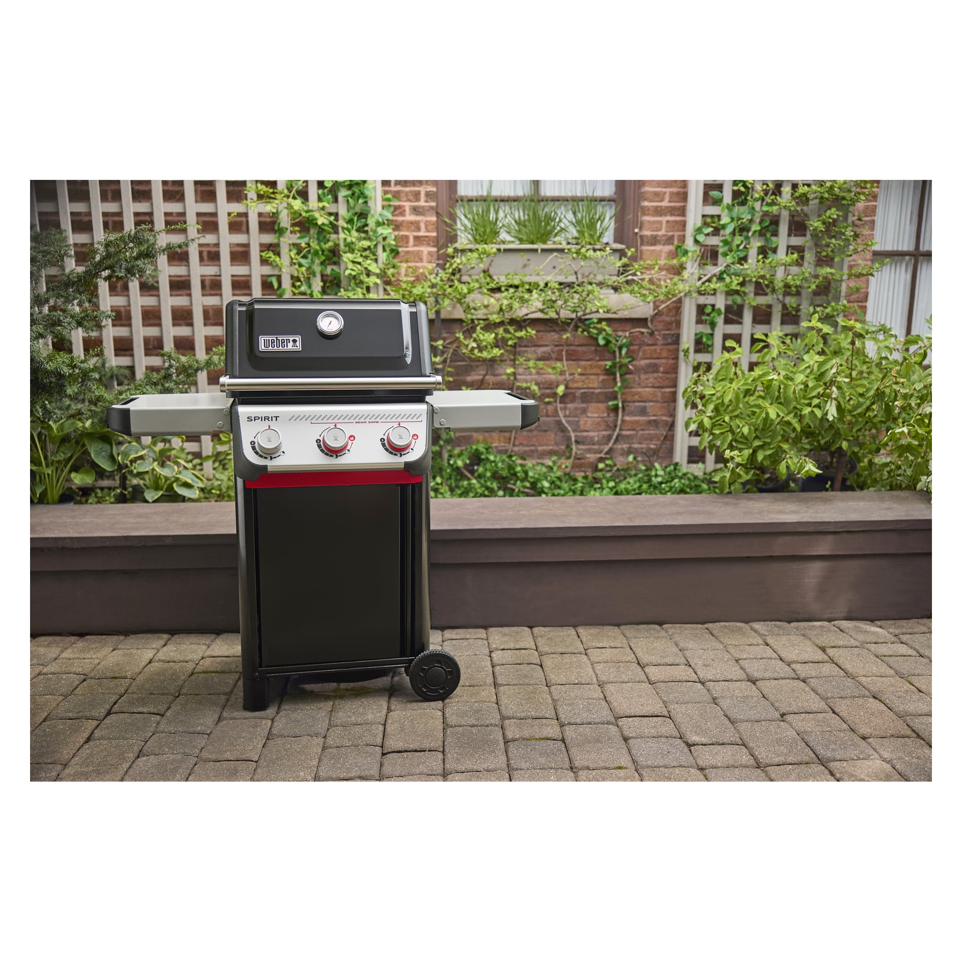 En stängd svart trebrännargrill står på en stenlagd uteplats, med gröna växter och en tegelvägg med spaljéer bakom.