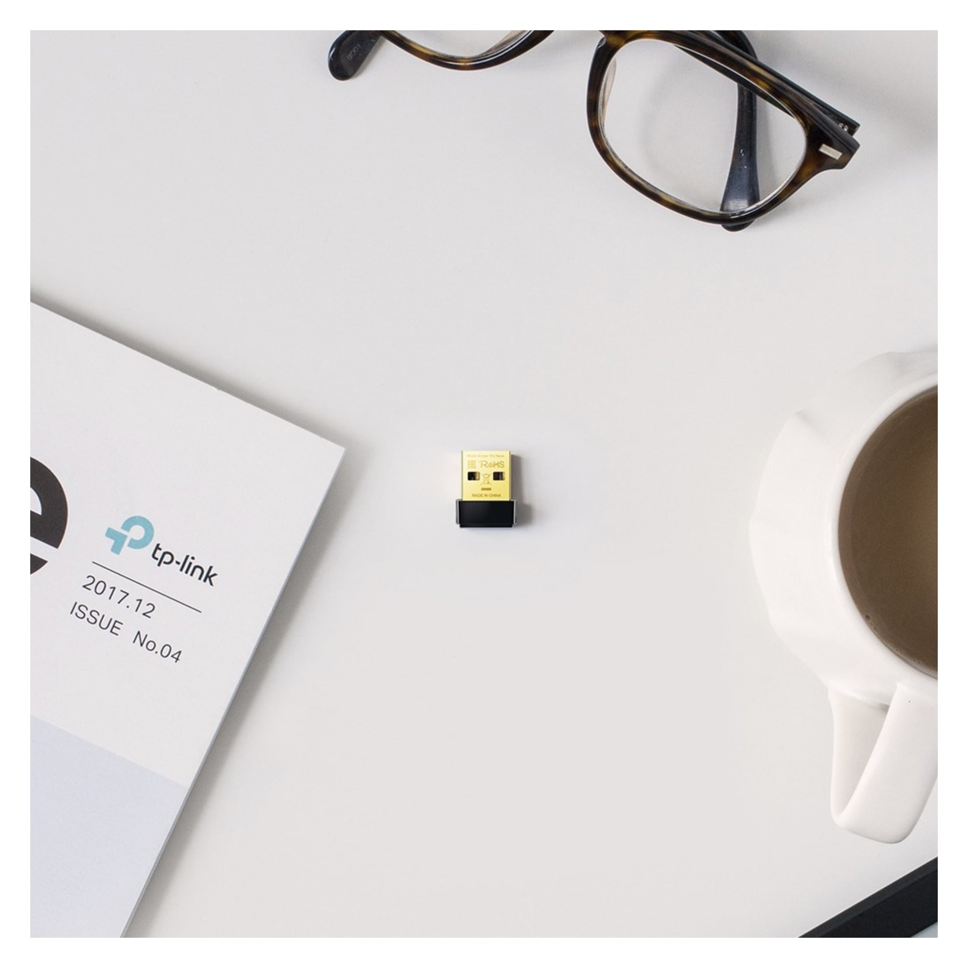 En guld- och svart USB-adapter vilar bredvid en tidning, glasögon och en kopp kaffe på ett vitt skrivbord.