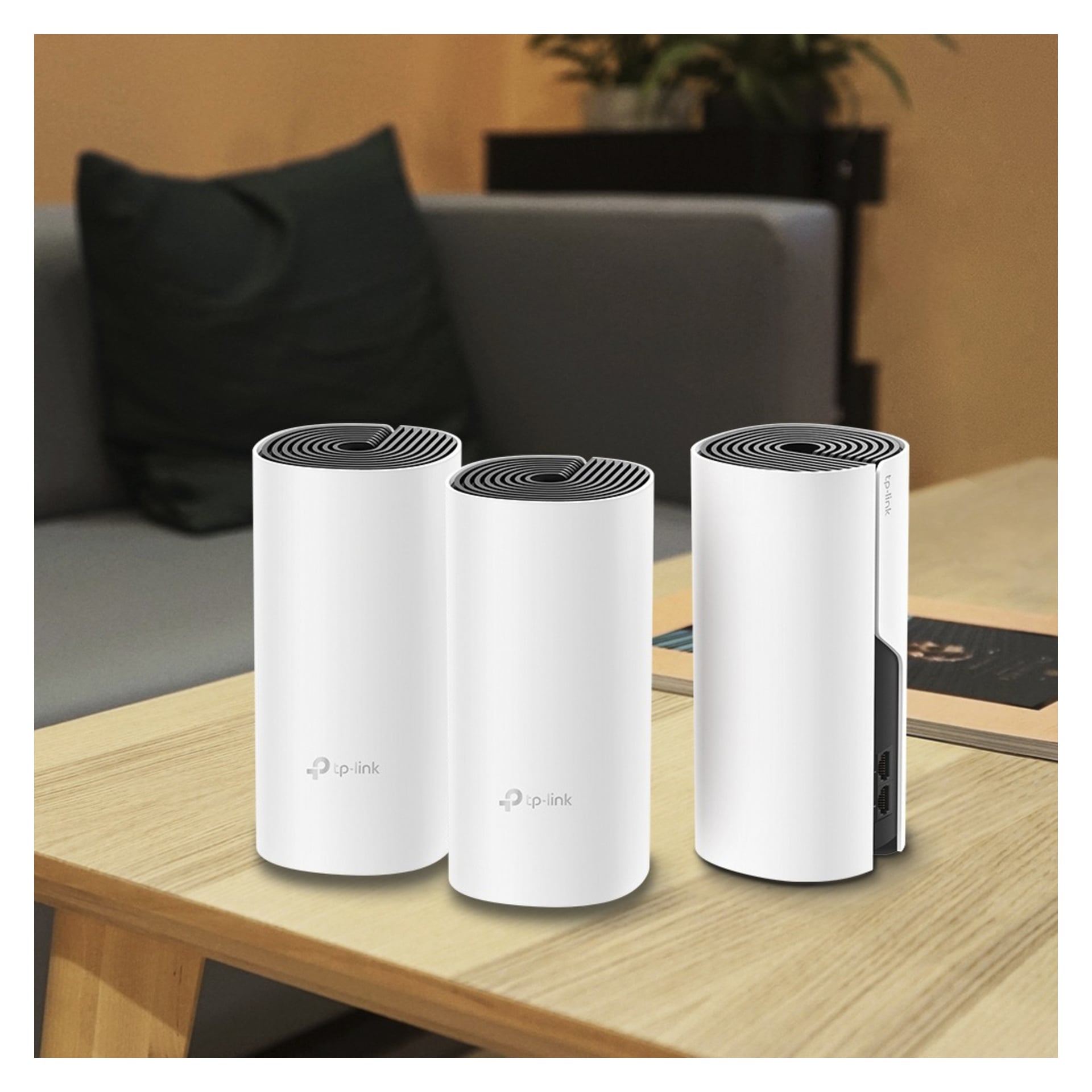 Tre vita cylindriska TP-Link-enheter med övre ventiler står på ett lätt träbord i ett vardagsrum med en grå soffa och böcker bakom.