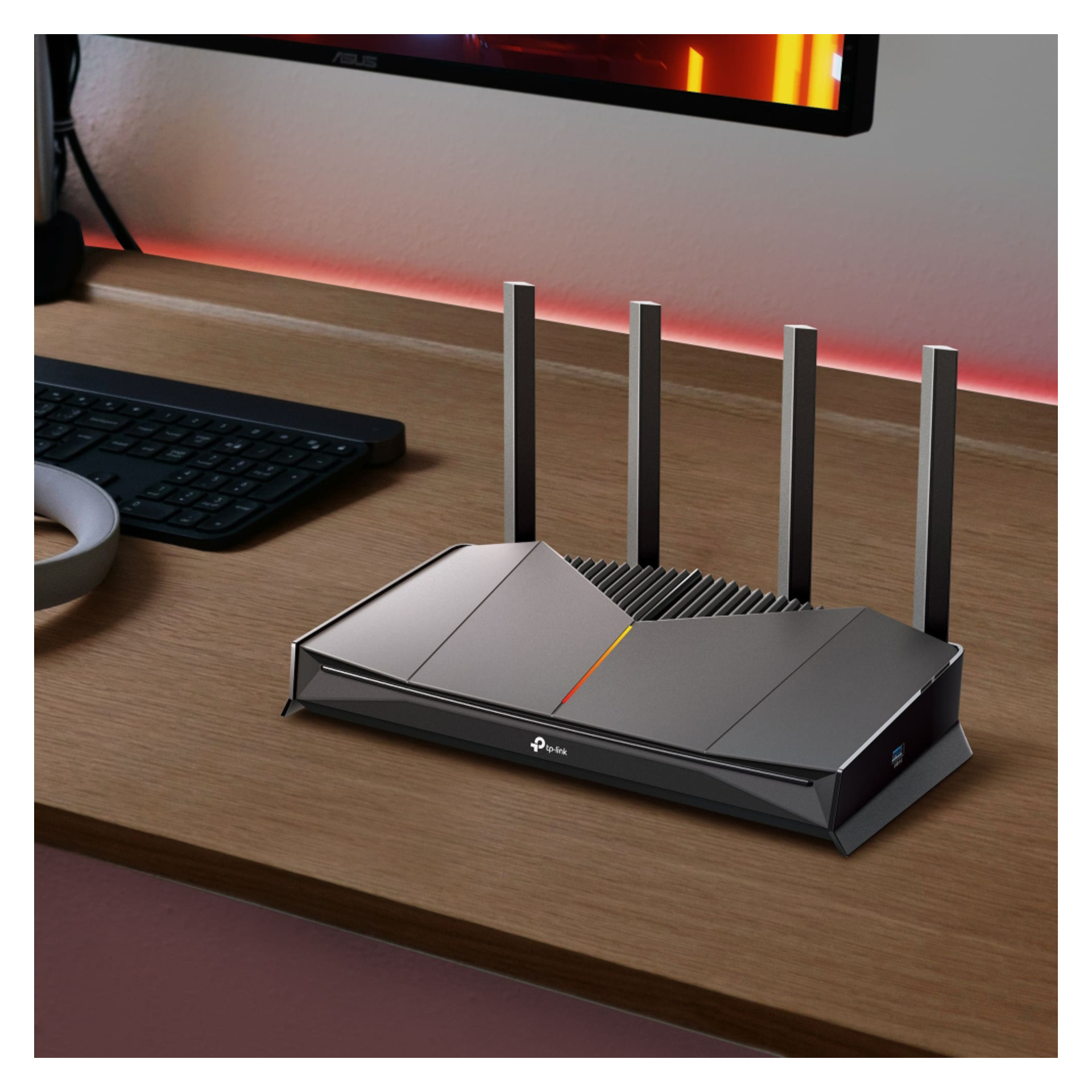 En trådlös router med fyra antenner placeras på ett träbord bredvid ett tangentbord och en skärm i ett hemmakontor.