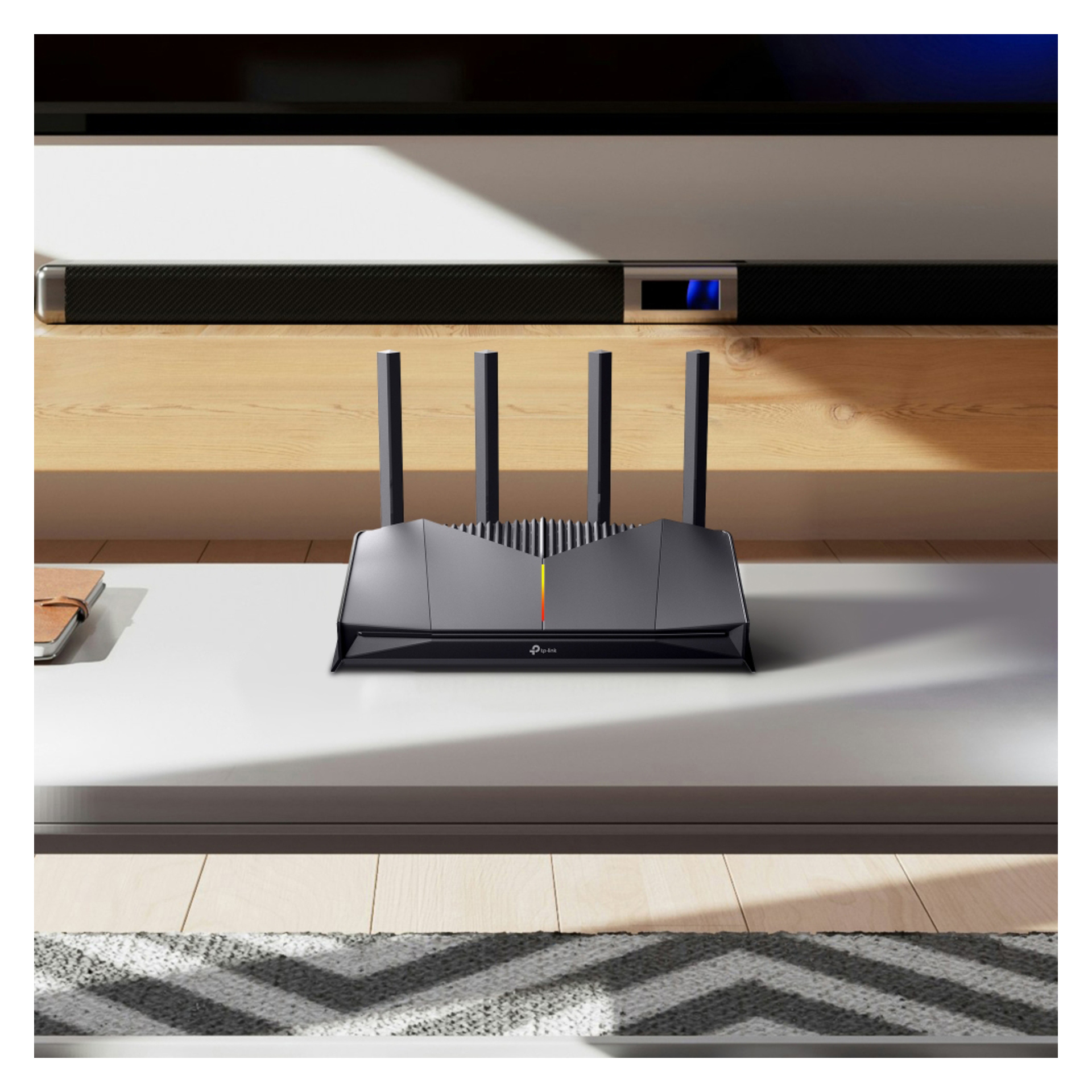 En svart trådlös router med fyra antenner står på ett vitt bord i ett modernt vardagsrum, med TV och soundbar på hyllorna bakom.