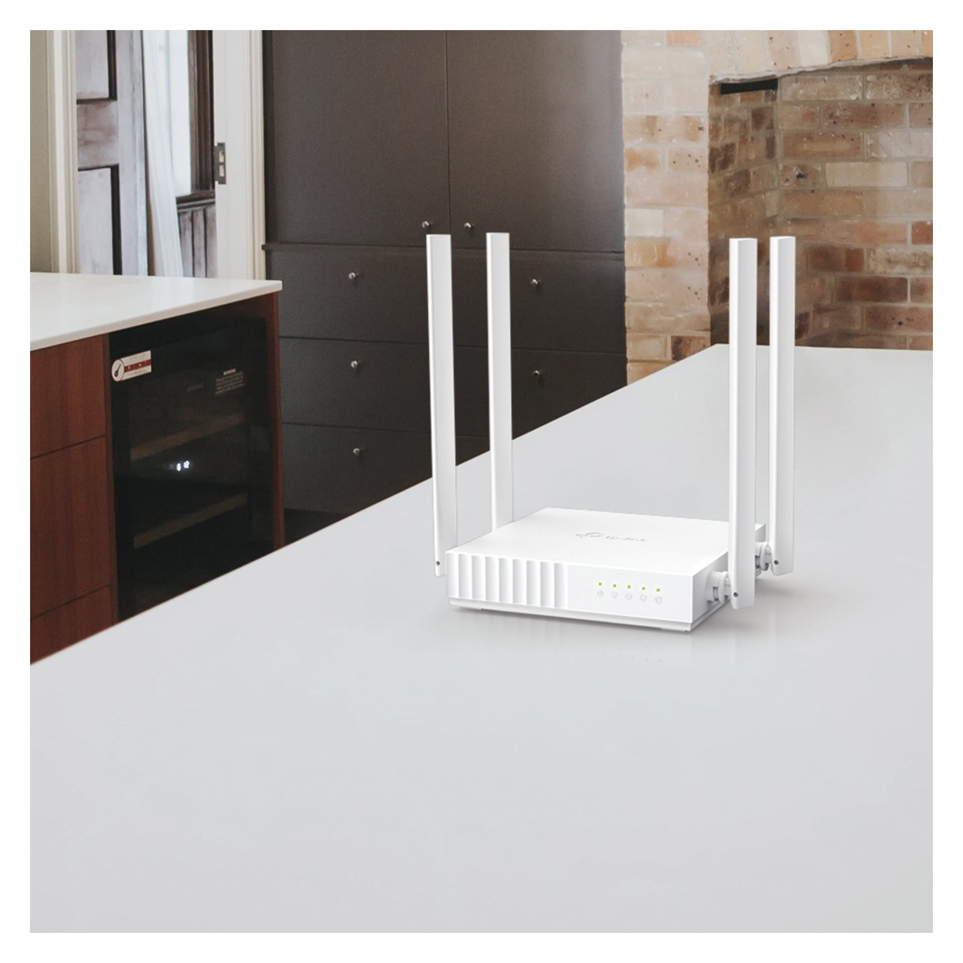 En vit trådlös router med fyra upprättstående antenner står på en ljus bänkskiva i ett modernt kök med tegel- och träskåp.