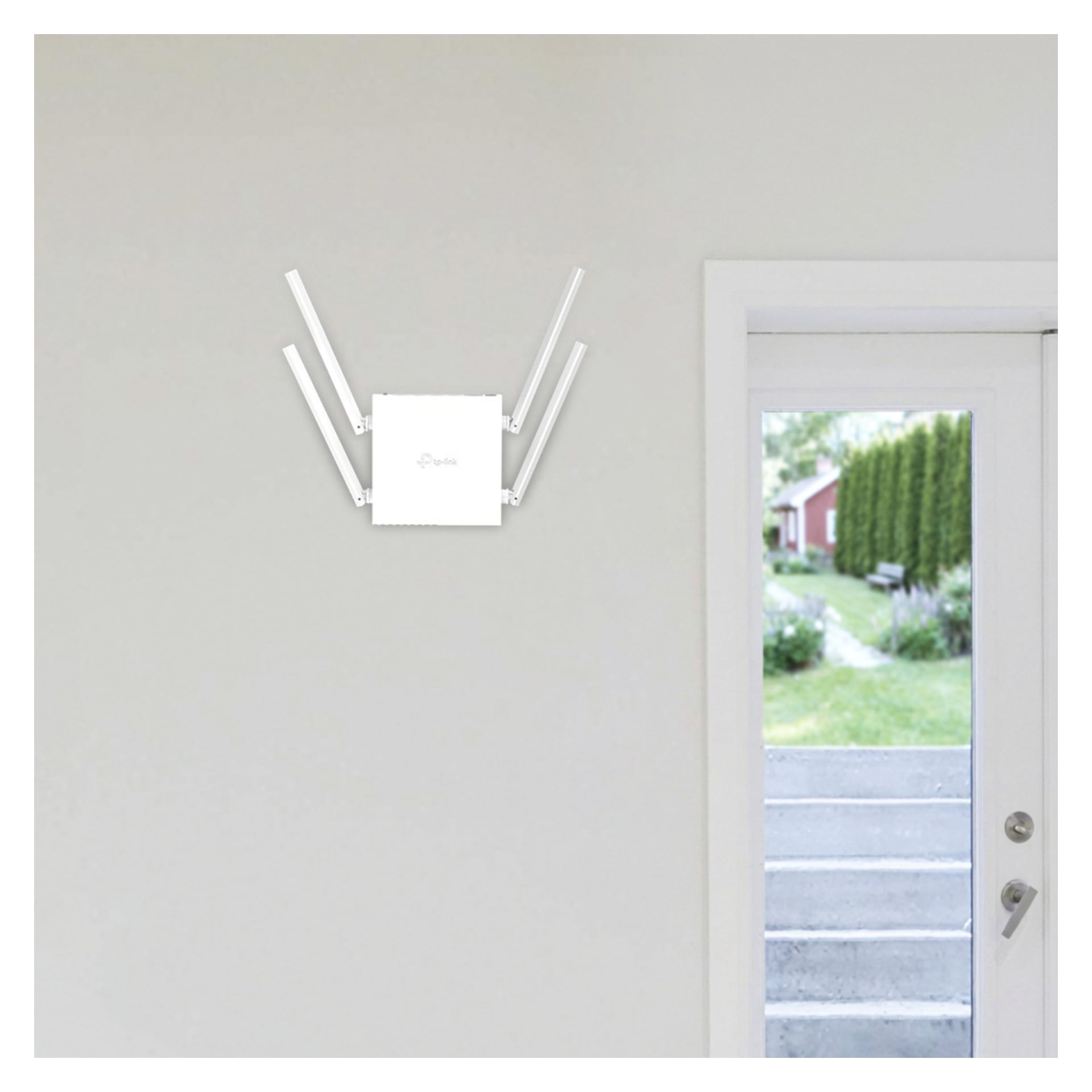 En vit trådlös router med fyra antenner är monterad på en ljusvägg bredvid en dörr med ett fönster som visar trappor och grönska utanför.