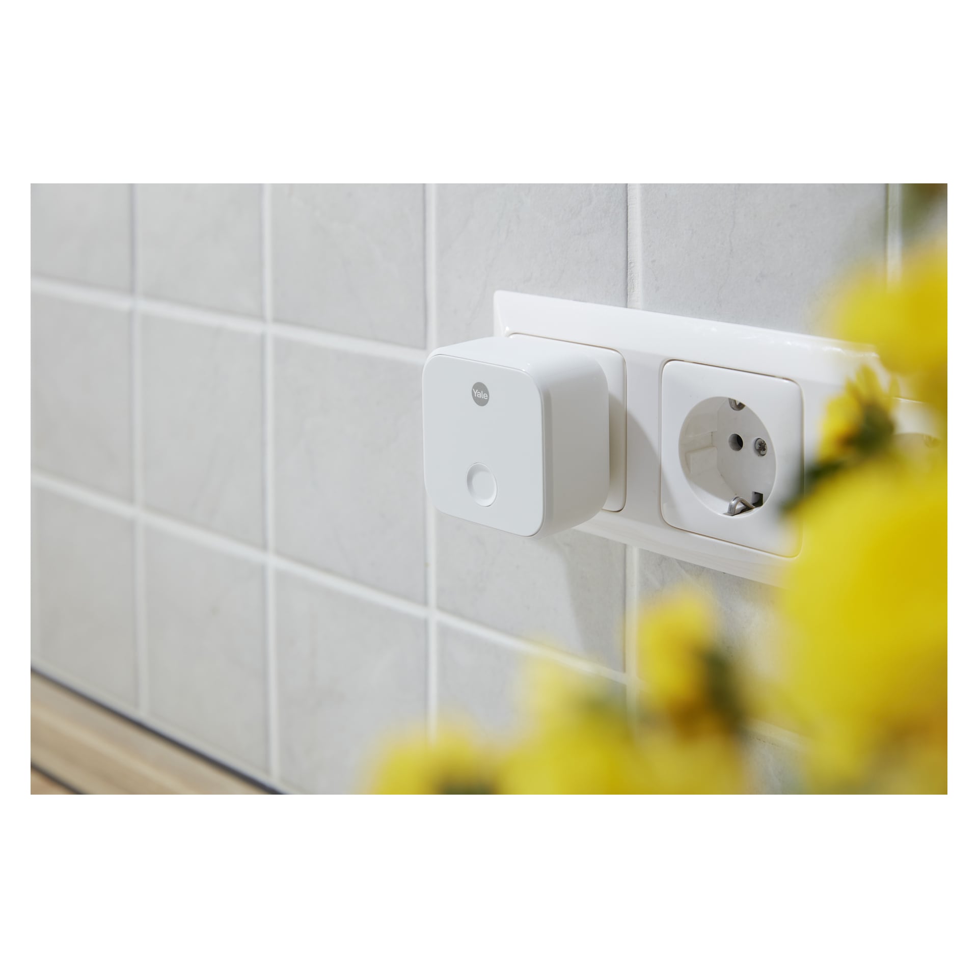 En hvit smart plugg er koblet til en kjøkkenveggkontakt på en flislagt flisvegg, med en annen stikkontakt i nærheten og gule blomster synlige.