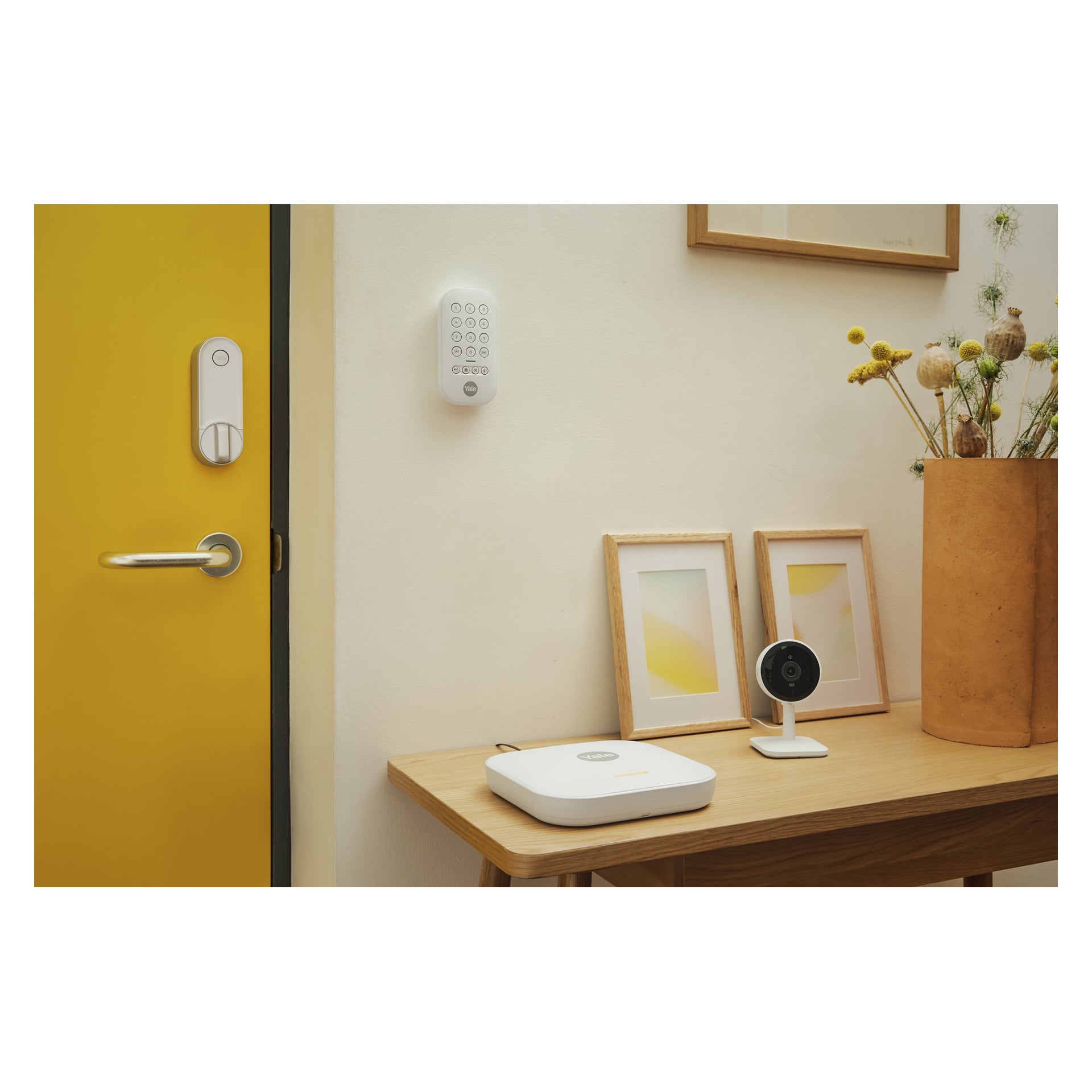 Inngangspartiet med gul dør, veggtastatur, trebord med smartenhet og sikkerhetskamera, innrammede trykk og høy blomstervase.