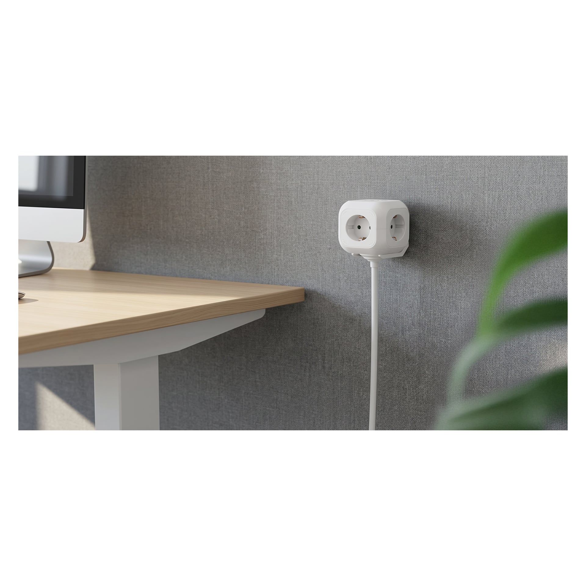 Ein weißer Würfel-Steckdosenleiste mit mehreren Steckdosen ist in eine Wand in der Nähe eines hellen Holzschreibtischs und Monitors angeschlossen, mit einer grünen Pflanze in der Nähe.