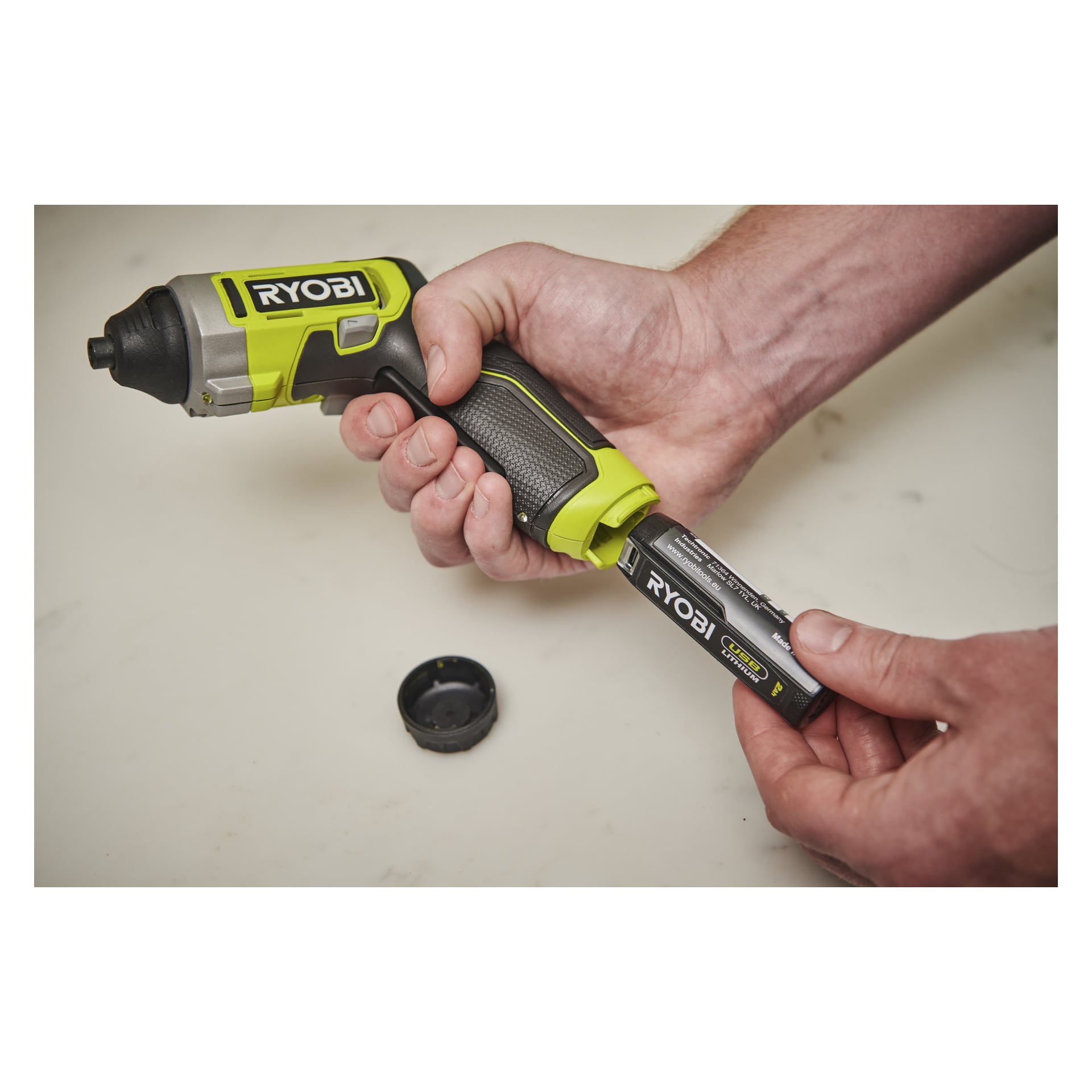 Någon placerar ett Ryobi-litiumbatteri i ett sladdlöst elverktygshandtag, med batterilocket vilande på en ljus yta i närheten.