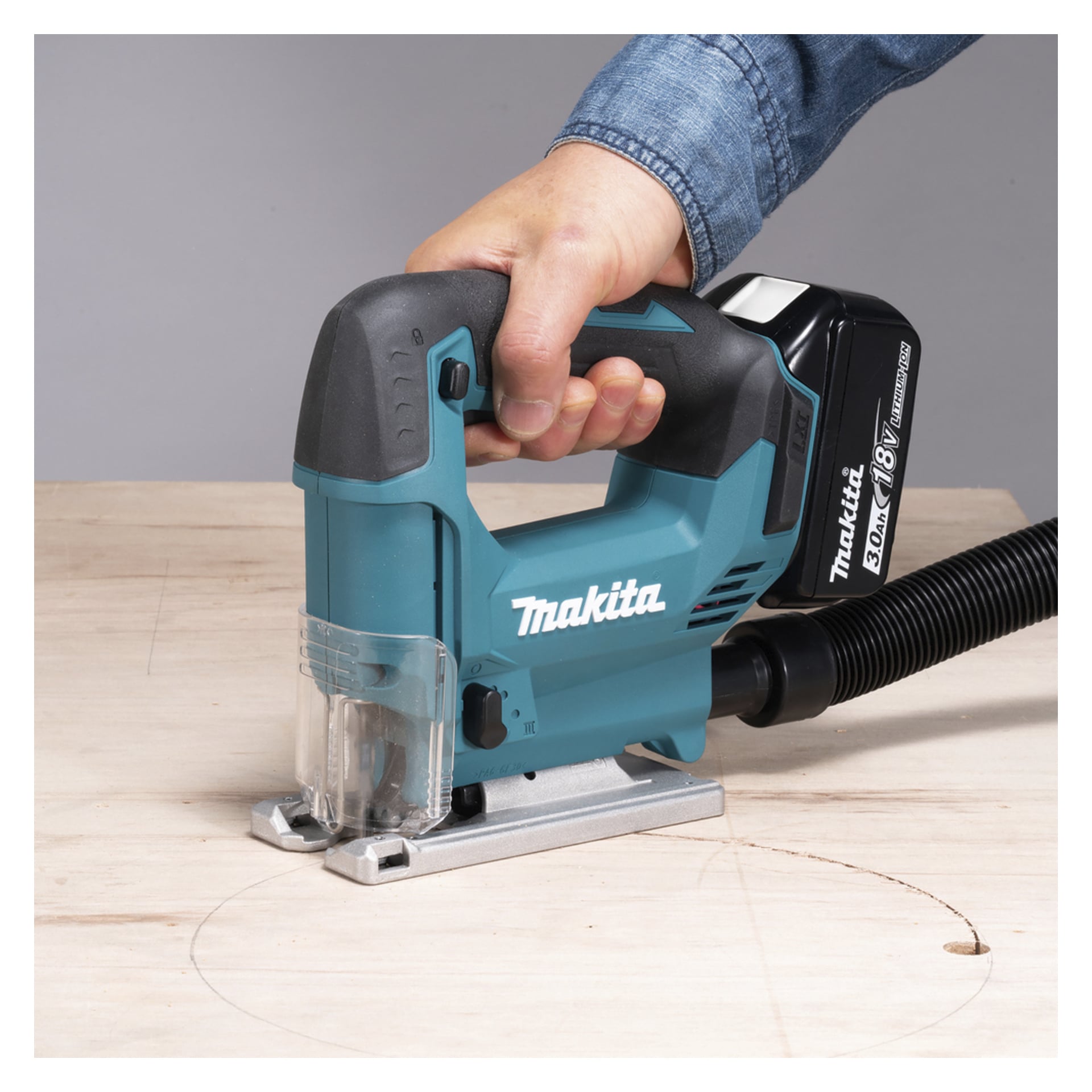 En person i blå skjorta använder en teal Makita sladdredodig sticksåg med svart batteri för att såga lätt trä på en plan yta.