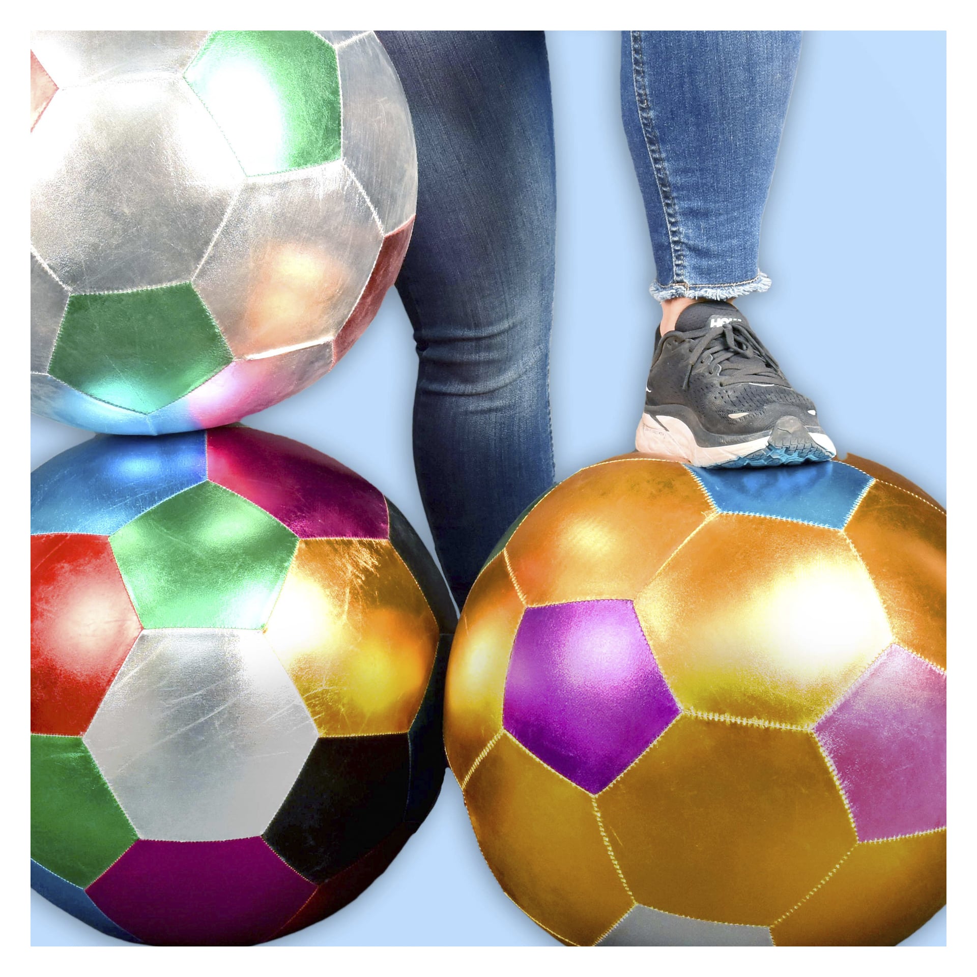 Iført jeans og grå joggesko hviler en person en fot på en gull- og lilla fotball. To metallkuler ligger i nærheten på blått.