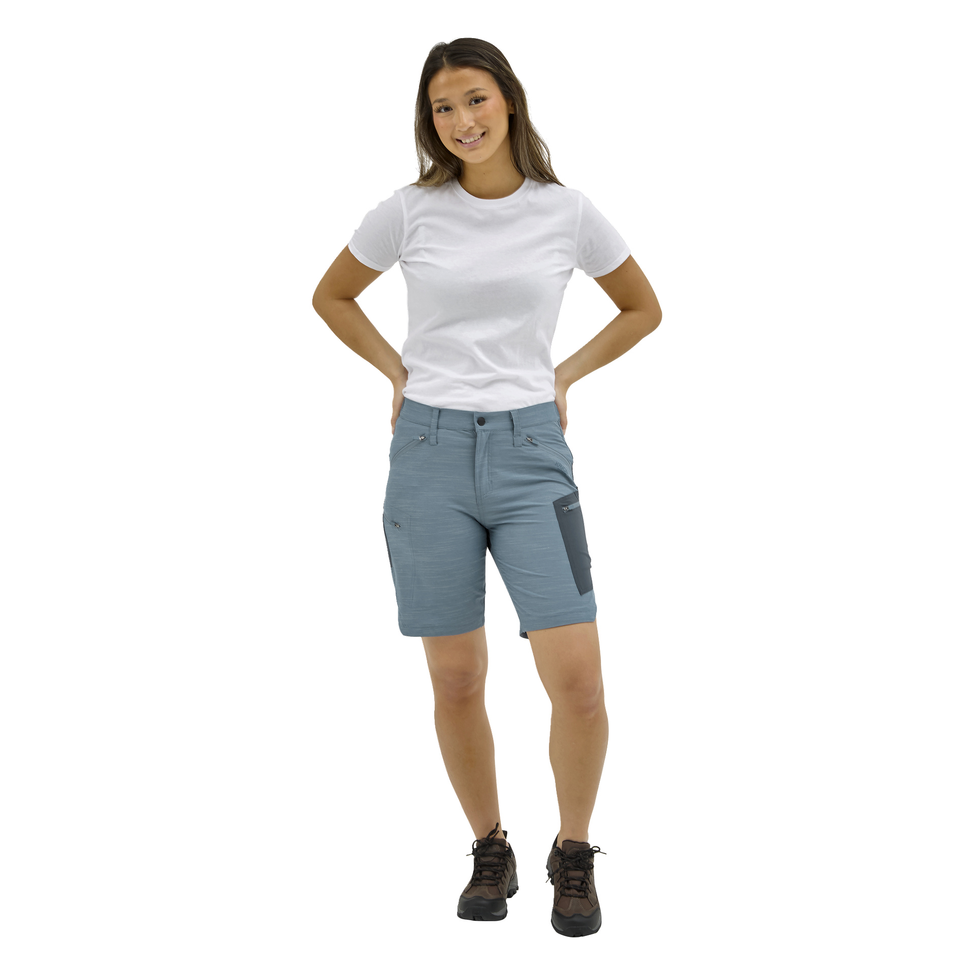 En kvinna vänder sig framåt, ler med händerna på höfterna, i en vit T-shirt, ljusblå knälånga shorts med dragkedjor och mörka vandringsskor.