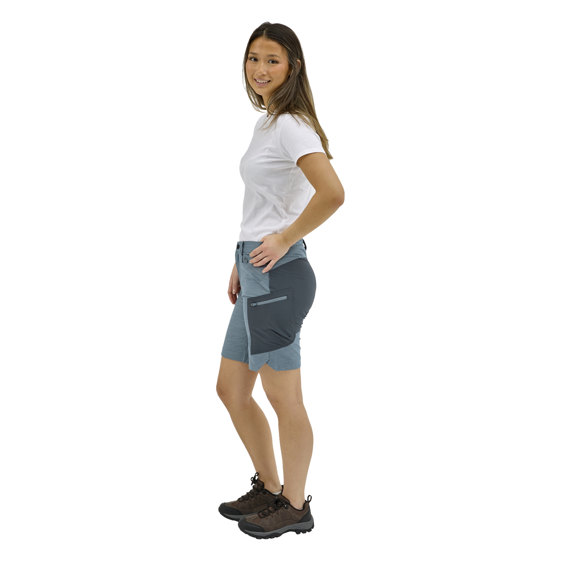 En kvinna står på sidan, leende, i en vit t-shirt, blå och grå shorts med fickor med dragkedja och bruna vandringsskor.