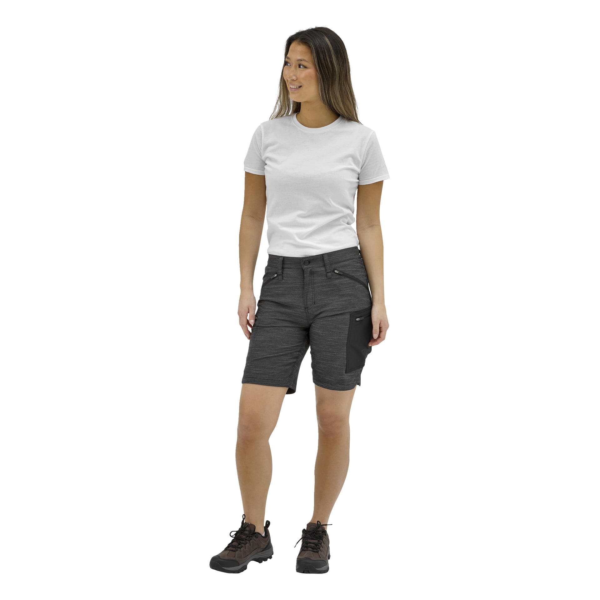 En kvinna står till vänster i en vit t-shirt, grå knälånga shorts med svart ficka och bruna vandringsskor, och ler milt.