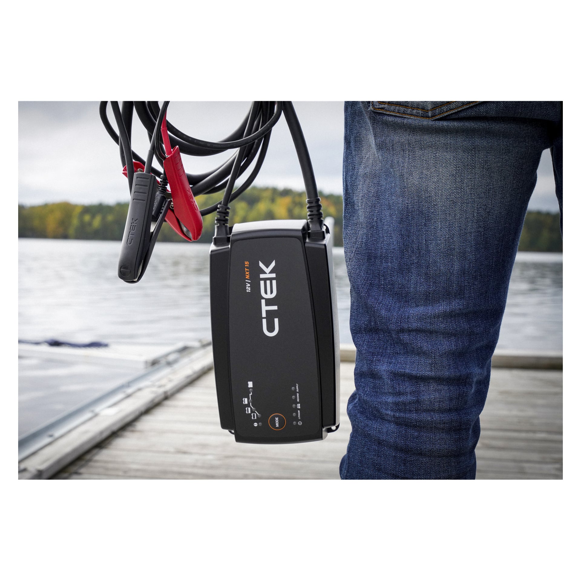 En person i jeans står på en brygga vid sjön och håller i en CTEK-batteriladdare med kablar; träd och vatten dyker upp bakom dem.
