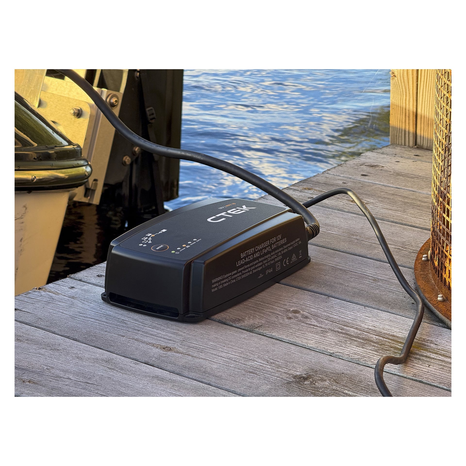 En svart CTEK-batteriladdare med kablar står på en träbrygga vid vattnet, solljus kastar skuggor och en metallstolpe till höger.