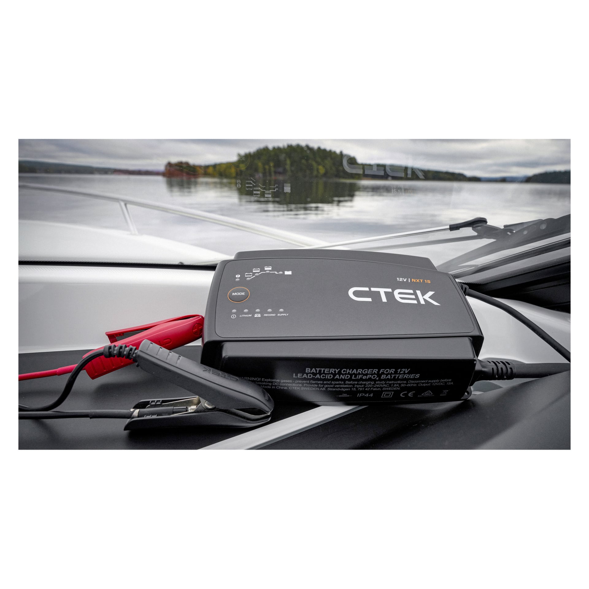 En CTEK-batteriladdare med startkablar vilar på båtens instrumentpanel, sjön och träd synliga utanför vindrutan.