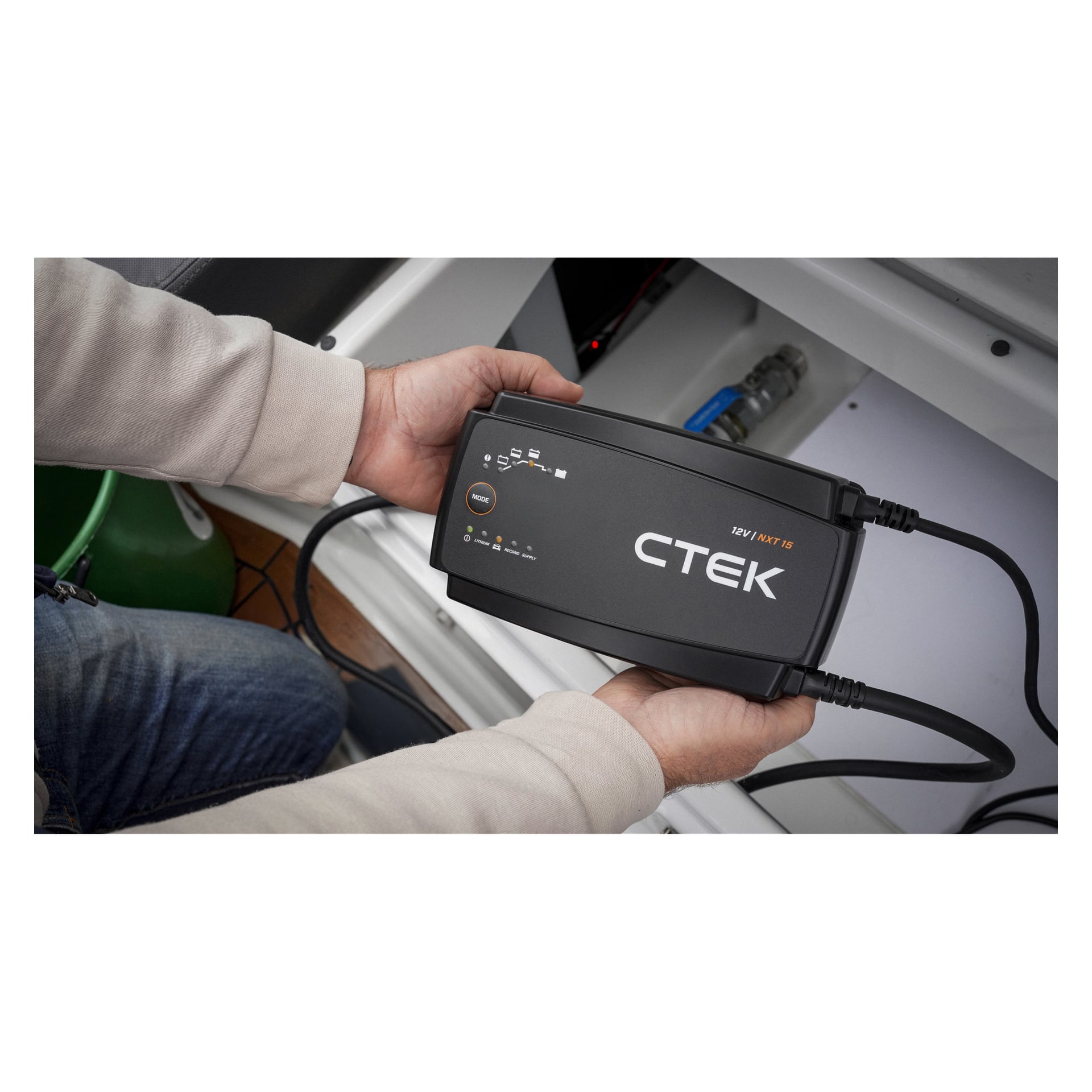 En person som bär en beige långärmad tröja och blå jeans håller en CTEK 12V batteriladdare med tända indikatorlampor i ett fack.