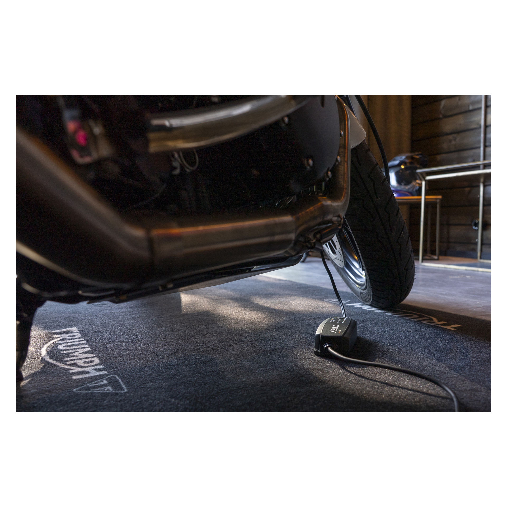 Zbliżenie na przednie koło i wydech motocykla, podłączona ładowarka baterii, stojąca na macie Triumph w środku.