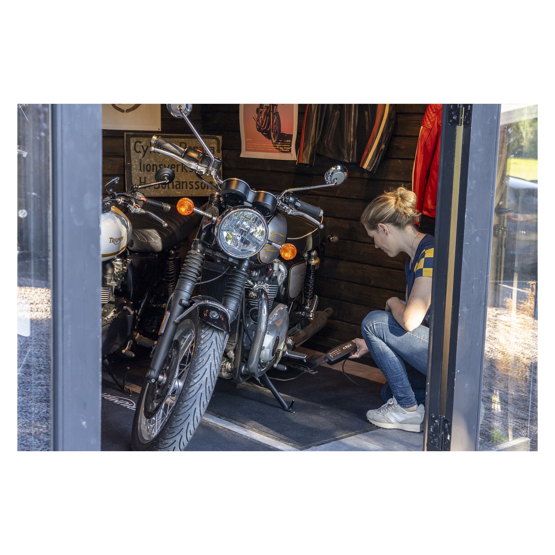 Ktoś klęczy przy motocyklu w garażu, używając przenośnego urządzenia przy silniku. Na ścianie za nim wisi kolejny rower i kurtki.