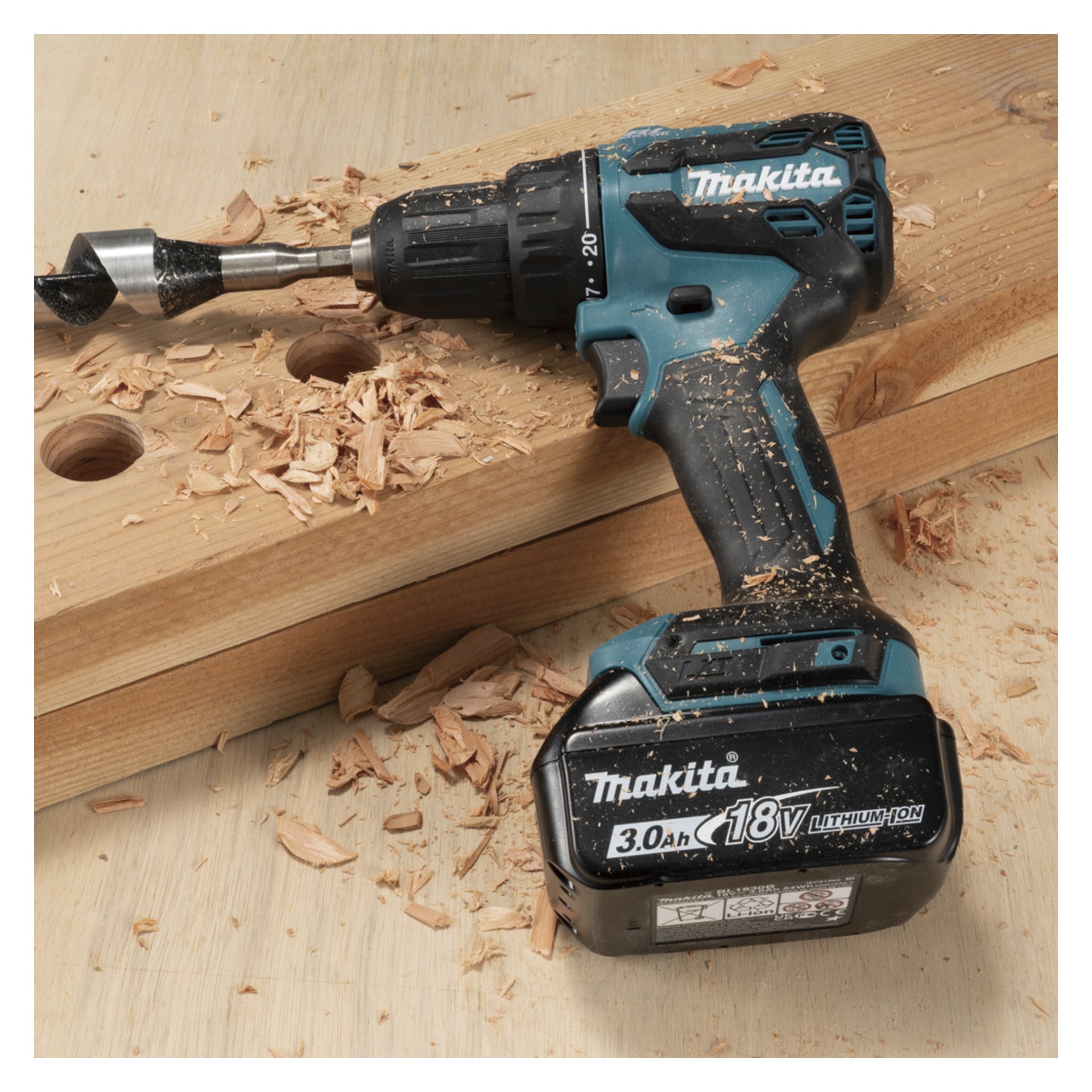 Makita 18V akkukäyttöinen porakone, jossa on iso terä, lepää puun päällä, johon on ripoteltu lastuja; Pinnassa näkyy kaksi suurta reikää.