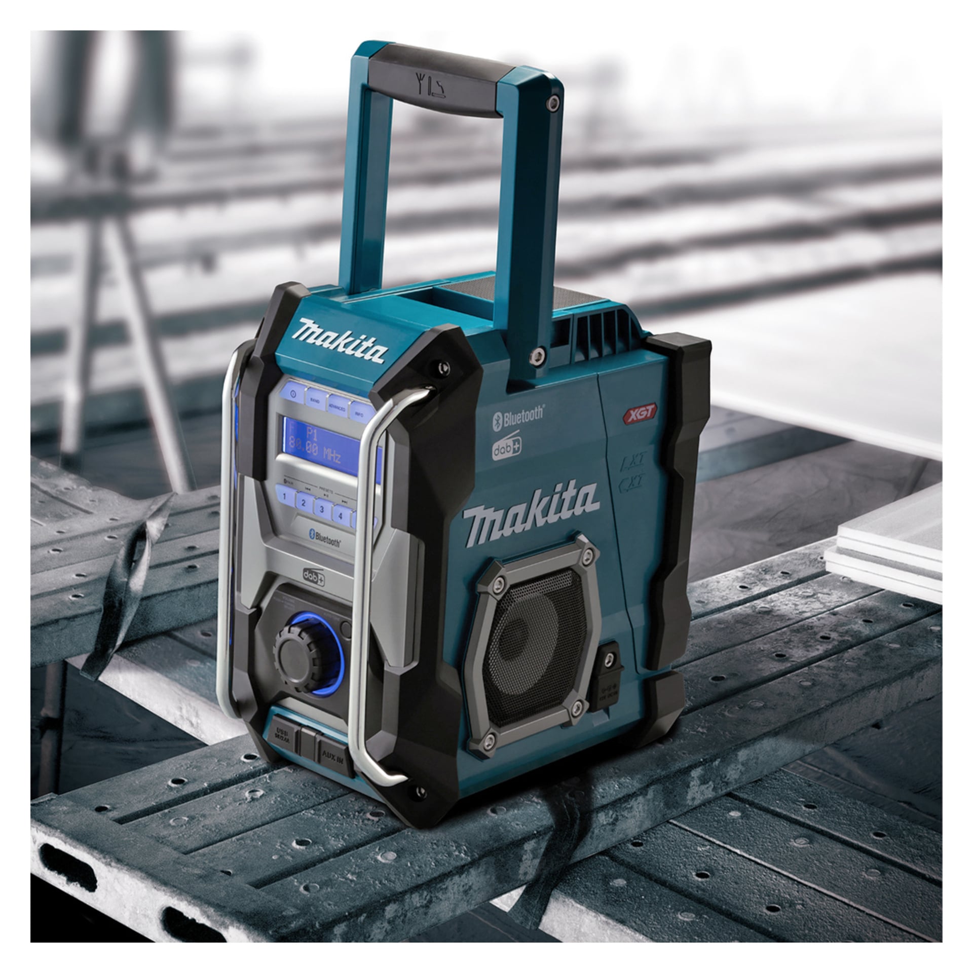 En blå och grå Makita arbetsplatsradio med Bluetooth och digital display vilar på en metallbänk, handtaget ovanpå, industriell bakgrund suddig.