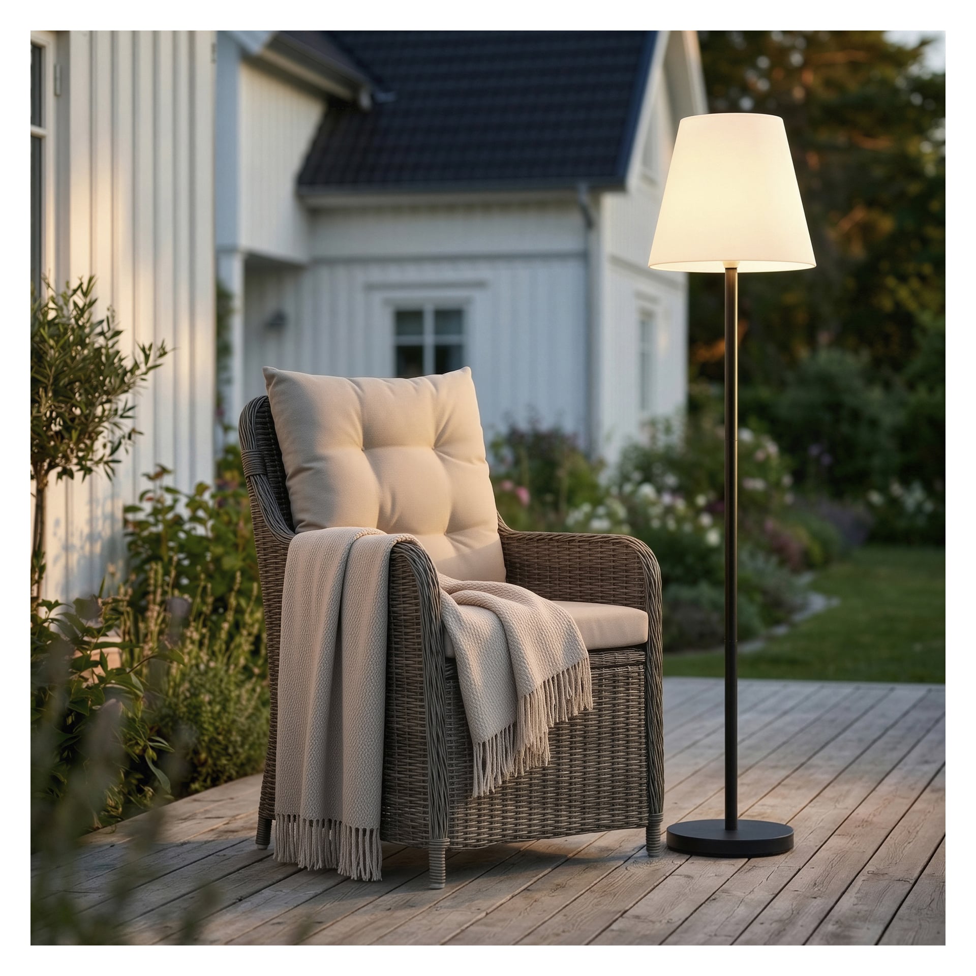 Ein Weidensessel mit beigefarbenem Kissen und einer Decke auf einer Holzterrasse neben einer brennenden Stehlampe, im Hintergrund ist der Garten zu sehen.