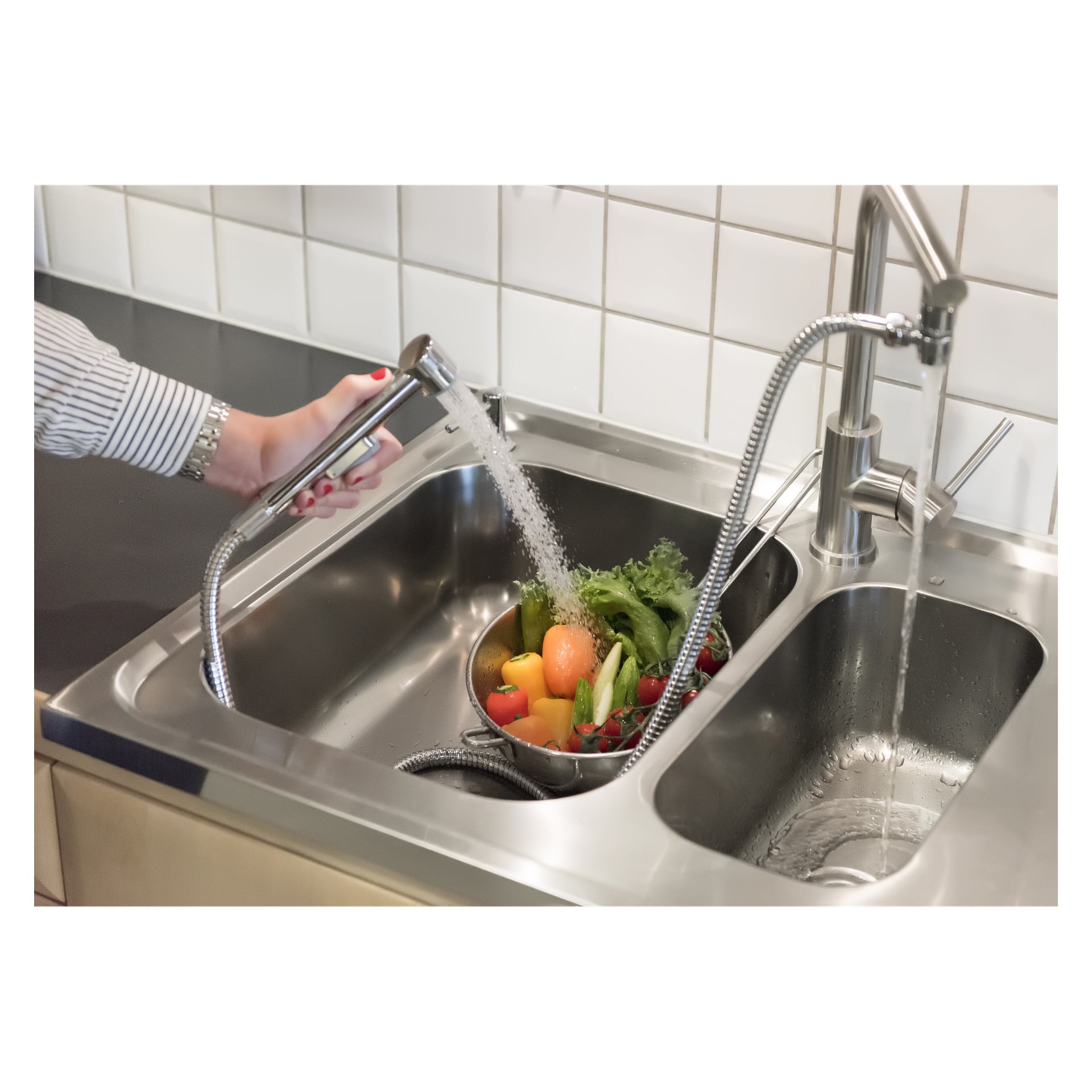 Någon sköljer en skål med blandade frukter och grönsaker under en avtagbar kranspruta i en diskho i rostfritt stål med klinkerskydd.
