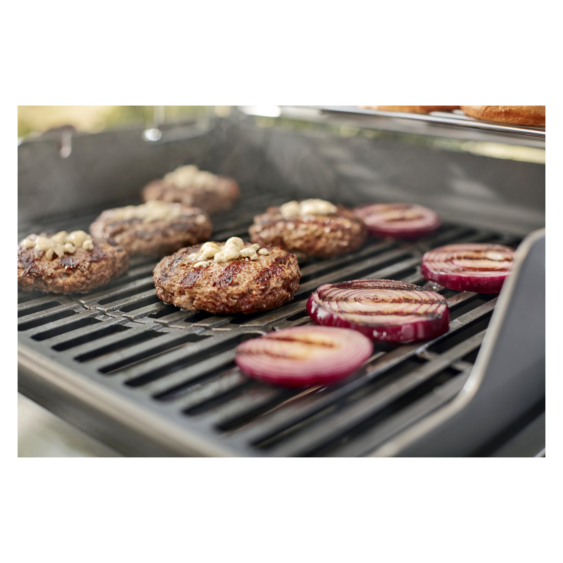 Burgerowe kotlety z roztopionym serem i grube plasterki cebuli skwierczą na gazowym grillu, gdy unosi się dym, a głębokie ślady po grillu tworzą się na zewnątrz.