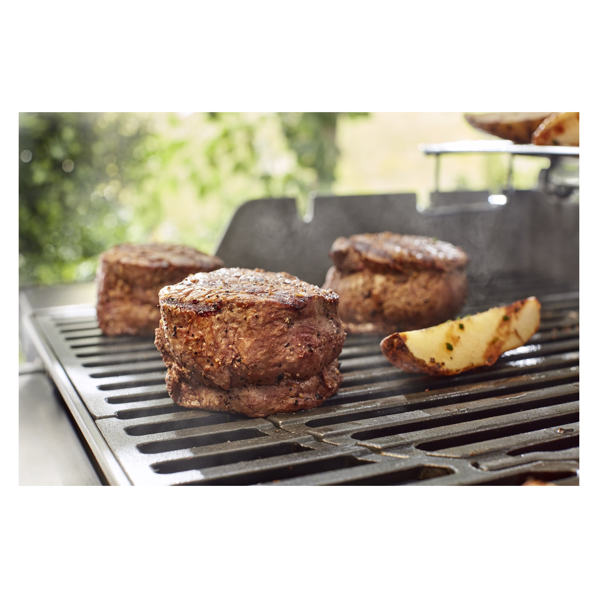 Trzy soczyste steki i przyprawiony kawałek ziemniaka skwierczą na gazowym grillu 8846, para unosi się, a za nimi rozmyta zieleń.