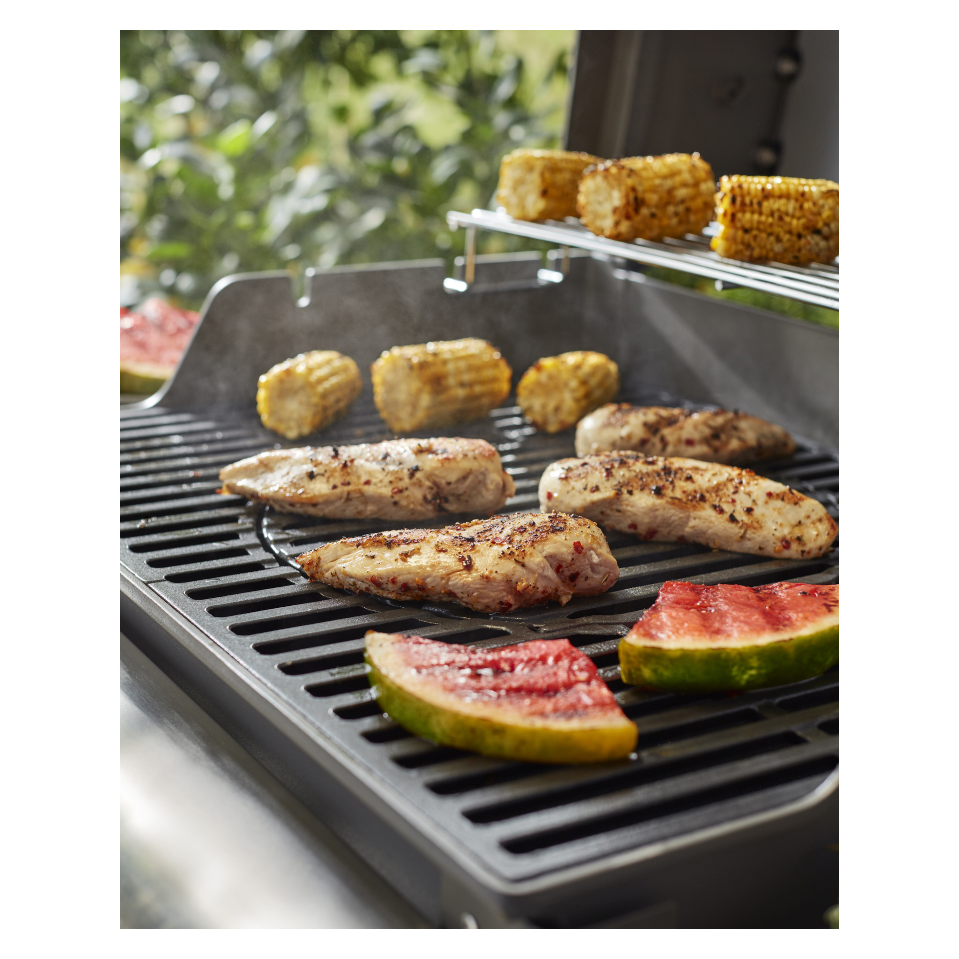 Kananrinnat, maissintähkä (osa yläritilällä) ja vesimeloniviipaleet grillataan kaasugrillillä; sumea vihreys takana.