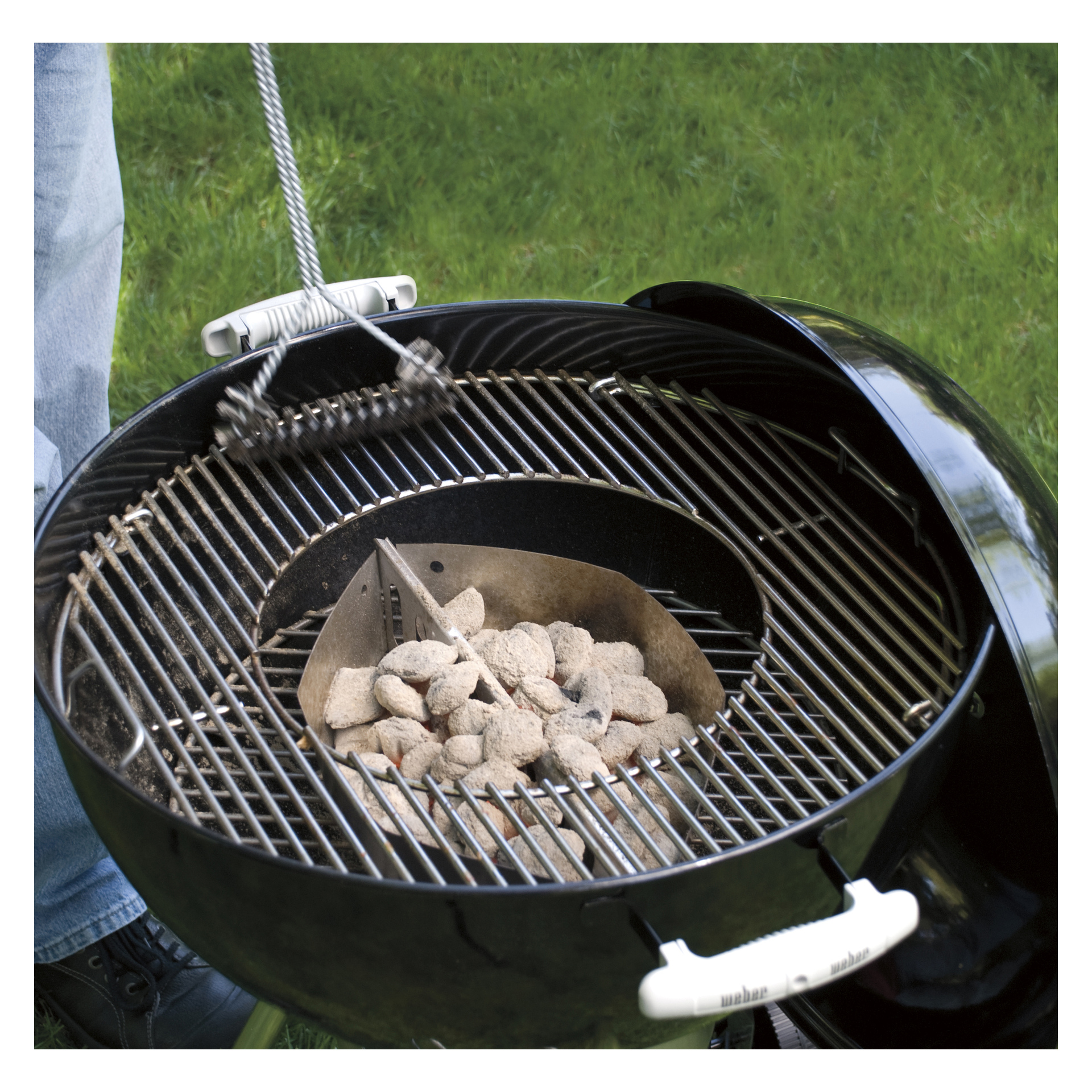 Någon skrubbar grillgallret på en kittelgrill 8835 med en stålborste; Obelysta briketter står i mitten på en gräsbevuxen gräsmatta.
