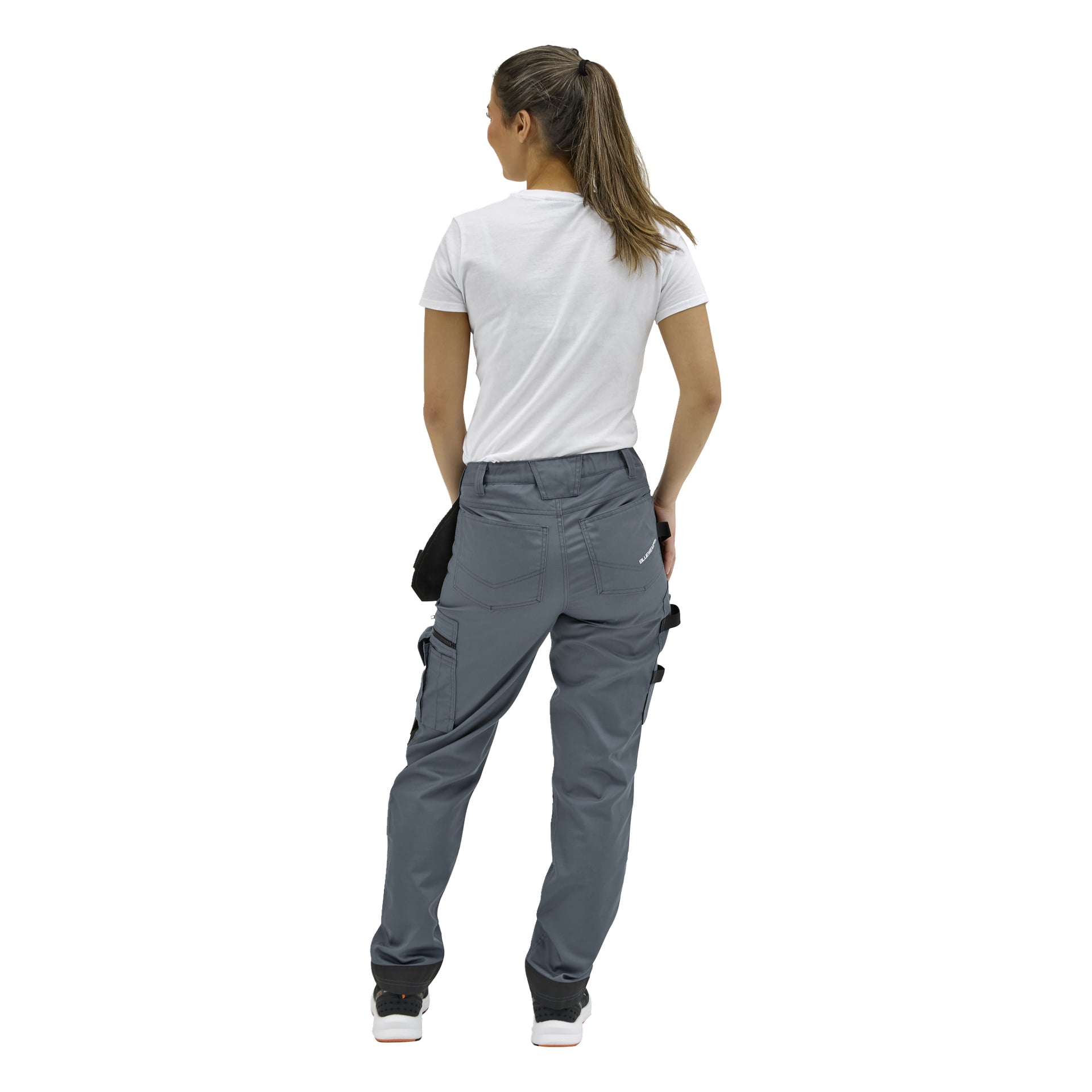 En person med hästsvans i vit t-shirt och grå verktygsbyxor står bort från kameran, handen i bakfickan.