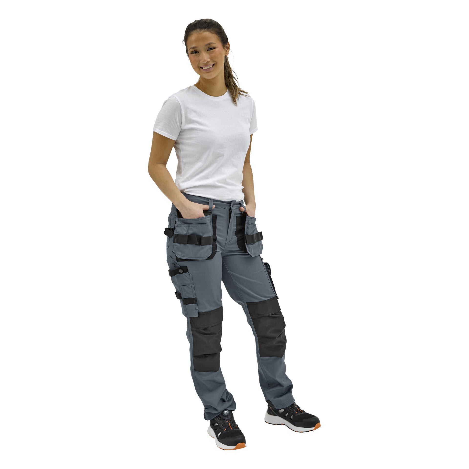 En leende person står med blicken framåt i en vit t-shirt, grå arbetsbyxor med svarta fickor och knälappar, samt svarta sneakers.