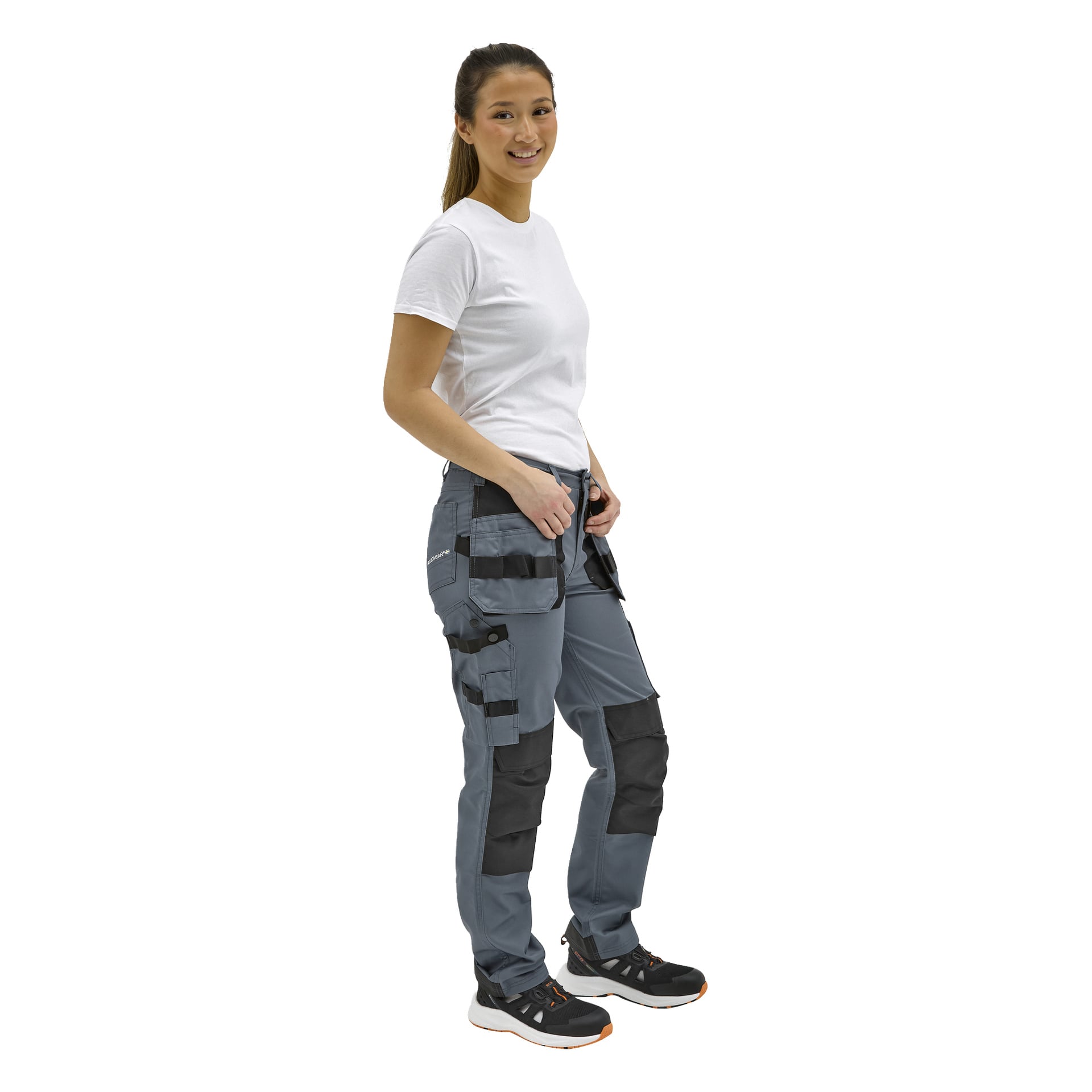 Leende kvinna i vit t-shirt, grå arbetsbyxor med svarta knälappar och fickor, svarta sneakers med vita sulor och orange.