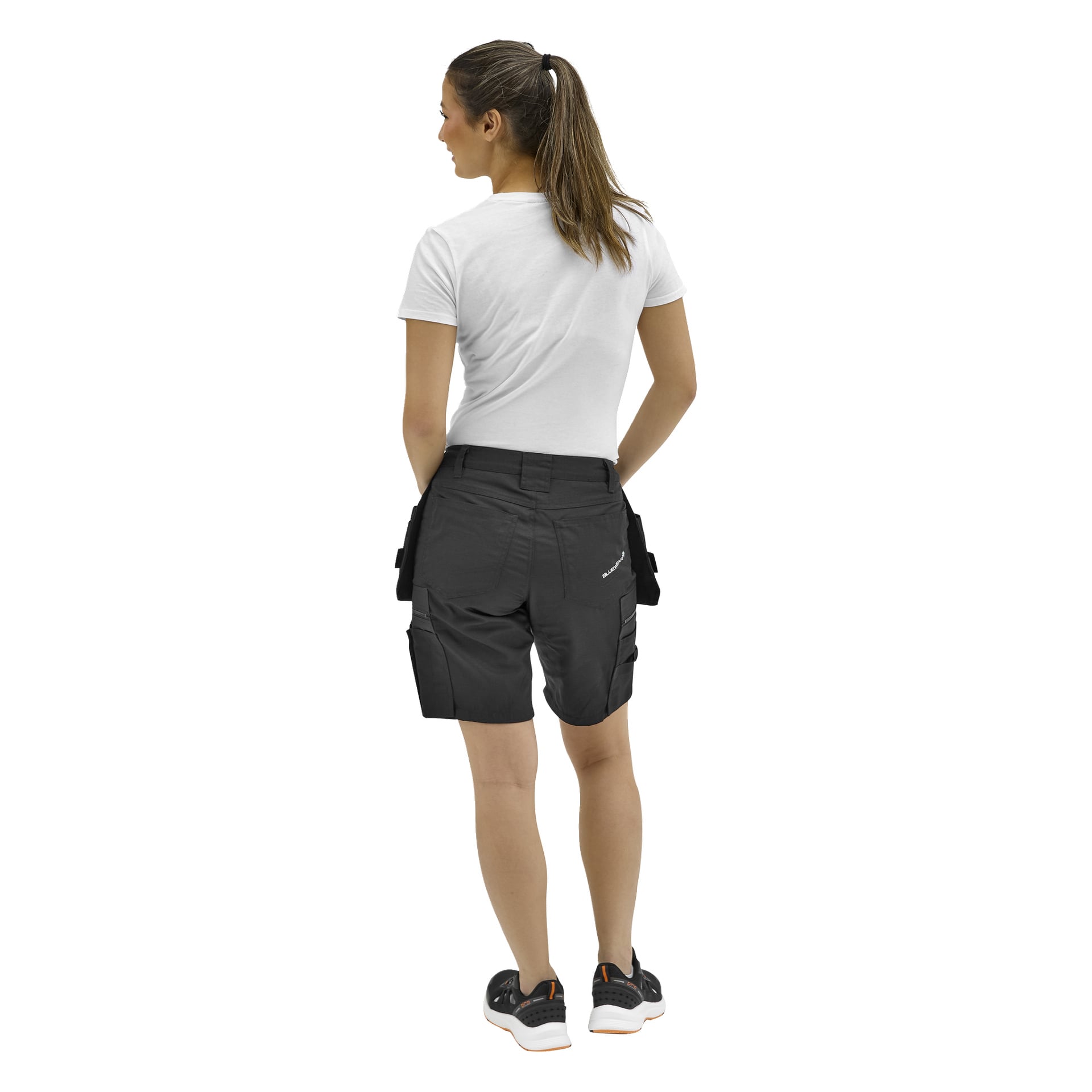 En kvinna med en lång brun hästsvans står med ryggen mot ryggen, klädd i en vit t-shirt, svarta arbetsshorts med fickor och svarta sneakers.