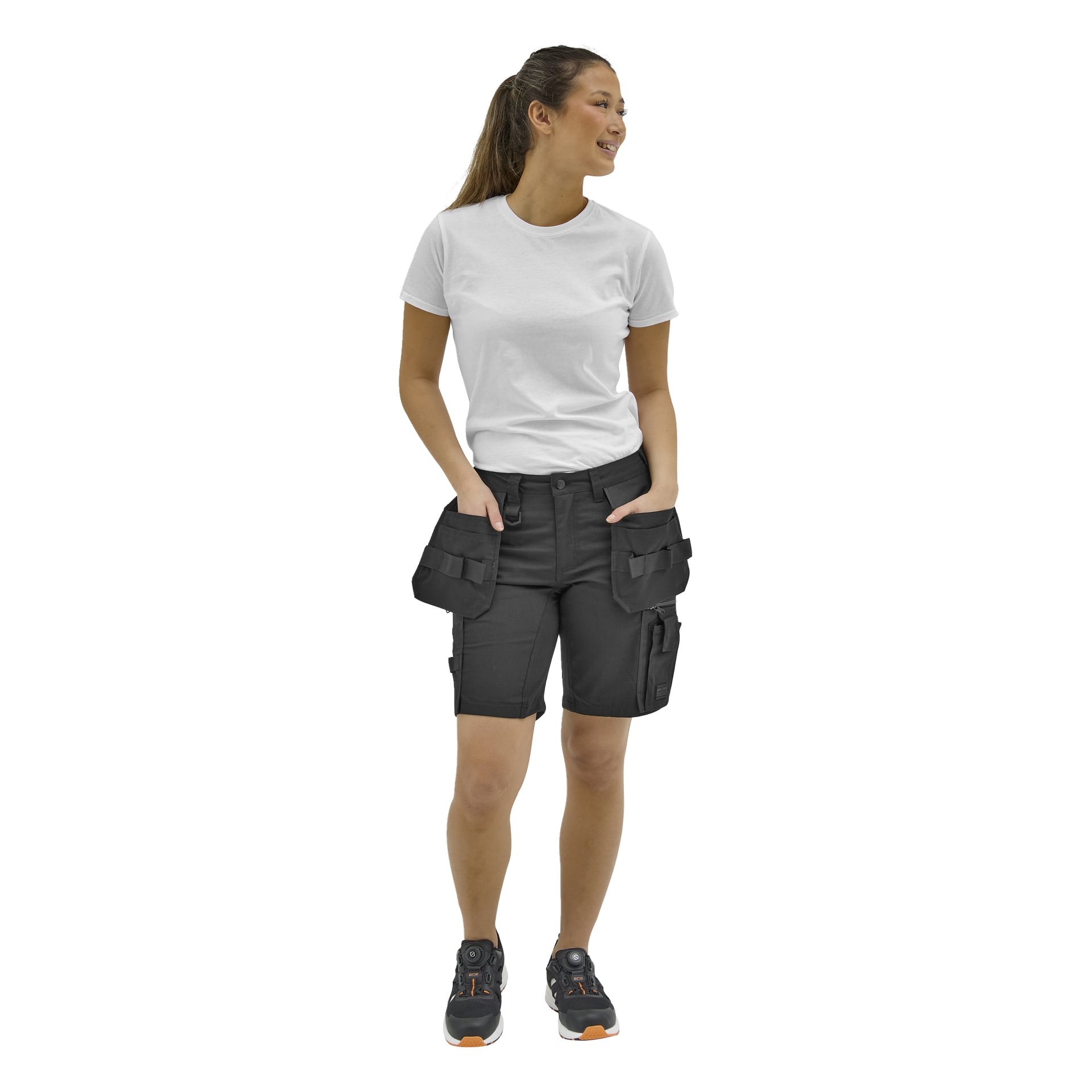 En person med hästsvans står i vinkel, klädd i en vit t-shirt, svarta cargoshorts och svarta sneakers med orange detaljer.