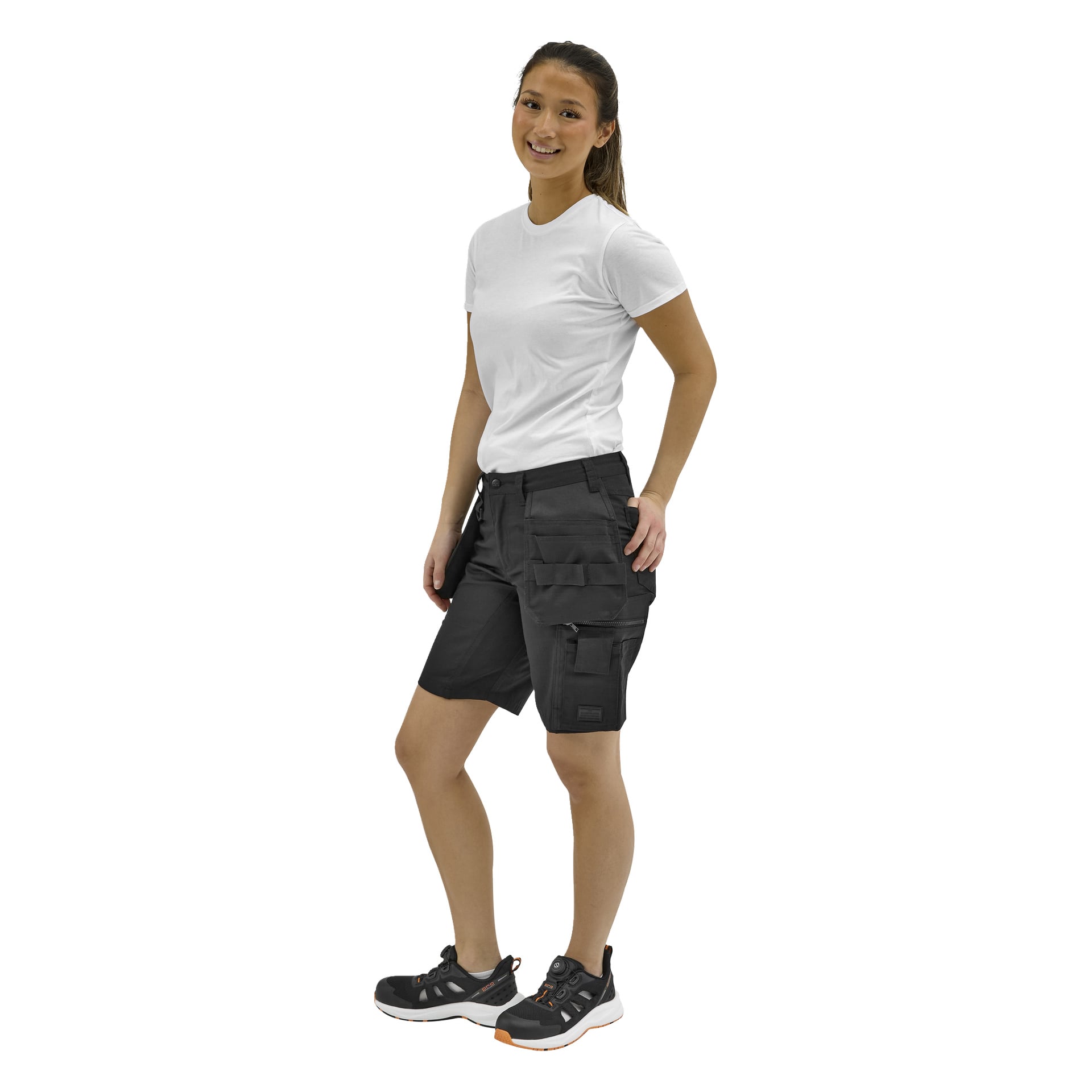 En leende kvinna står lätt vänd åt sidan, klädd i en vit t-shirt, svarta cargoshorts och svarta sneakers med orange detaljer.