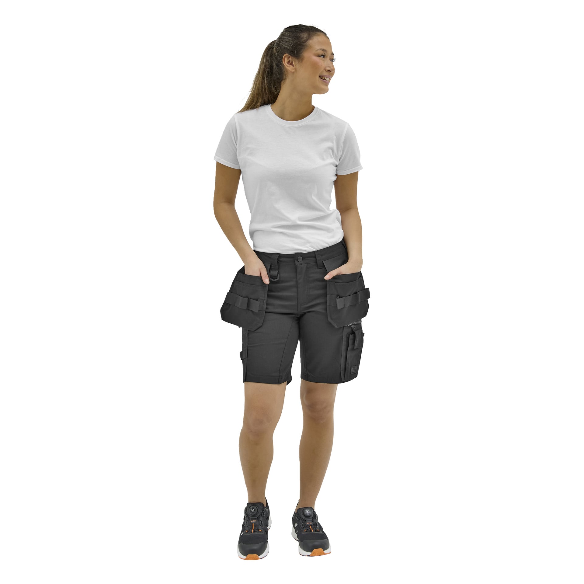 En person ler och tittar åt sidan, händerna i svarta cargoshorts, klädd i en vit t-shirt och svarta skor med orange detaljer.