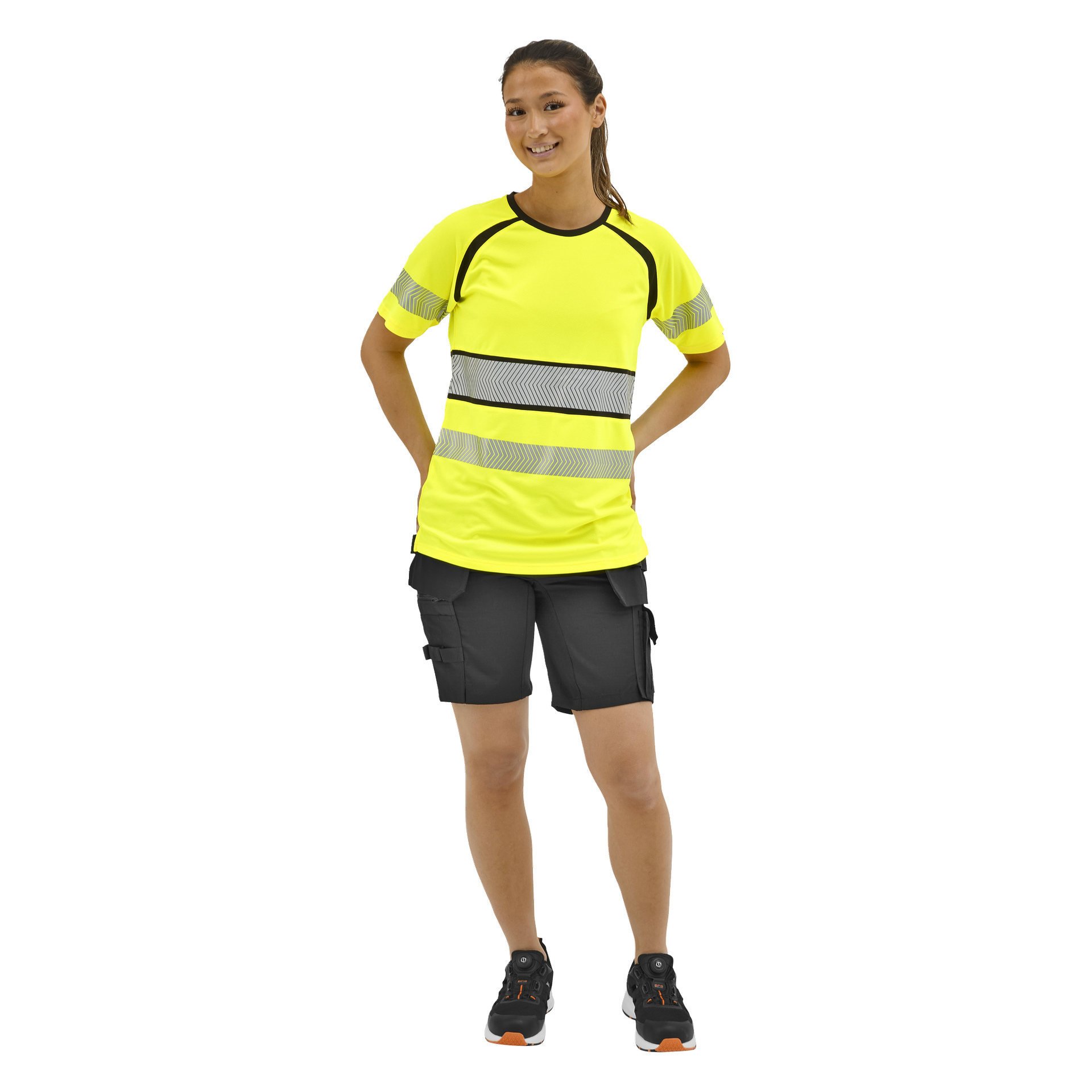 Eine Frau posiert mit den Händen in die Hüften in einem neongelben, reflektierenden Sicherheitsshirt, schwarzen Cargoshorts und schwarzen Turnschuhen mit orangefarbenen Details.