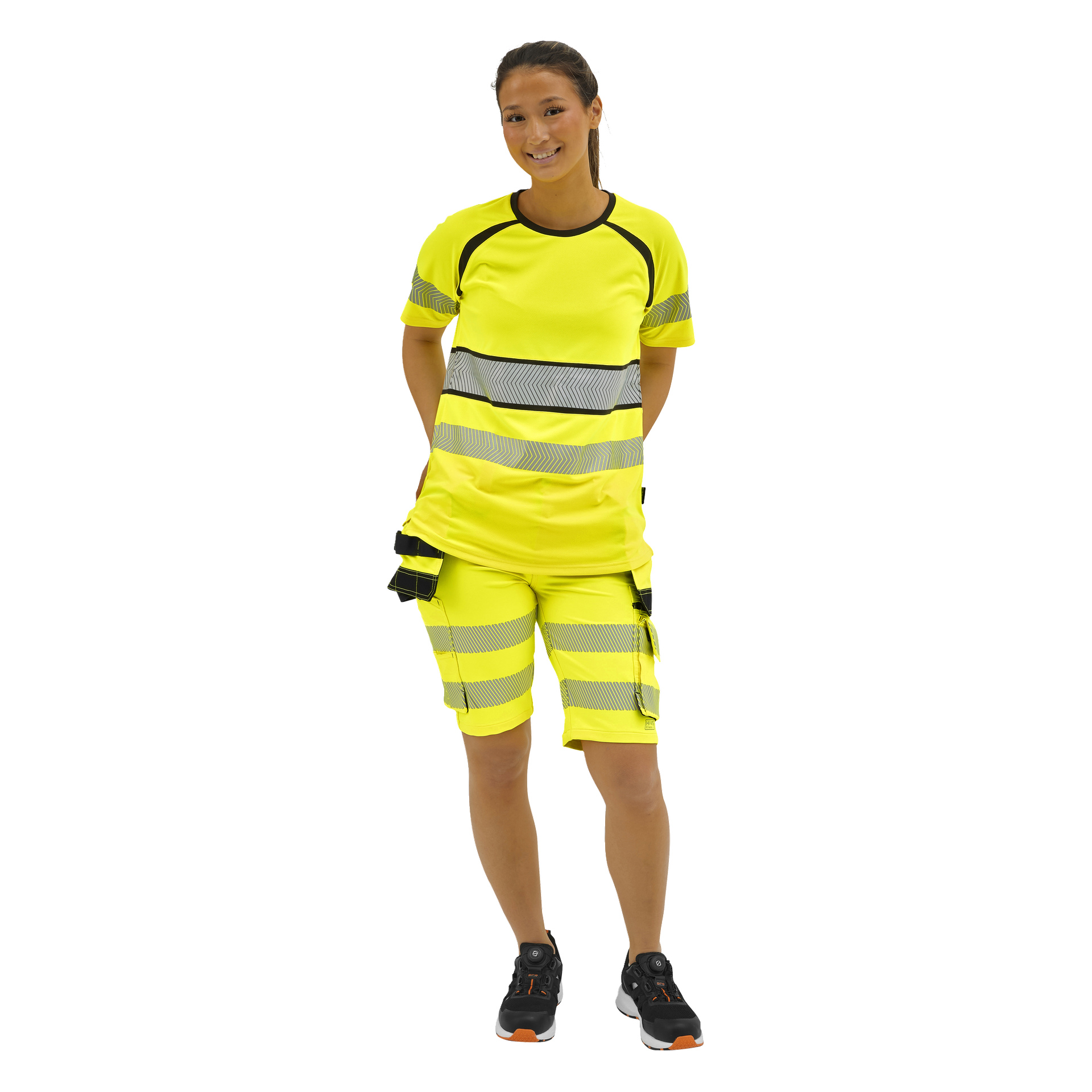 Eine lächelnde Person mit den Händen hinter dem Rücken in einem neongelben, passenden Set mit silbernen Streifen, schwarzen Akzenten und schwarzen Sportschuhen.