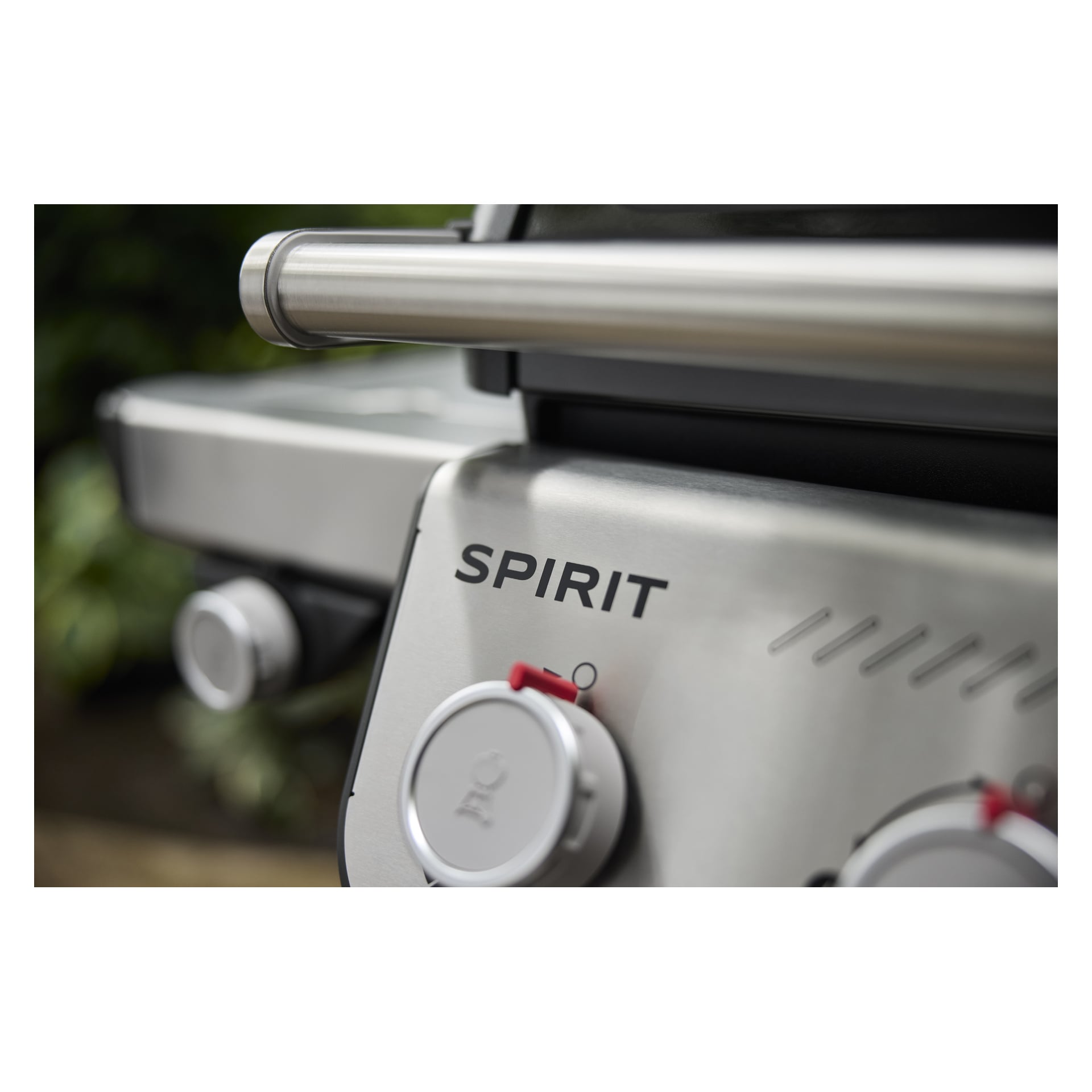 Kontrollpanelen på en rostfri Spirit gasgrill med två röda detaljer och handtag, grönska suddig i bakgrunden.