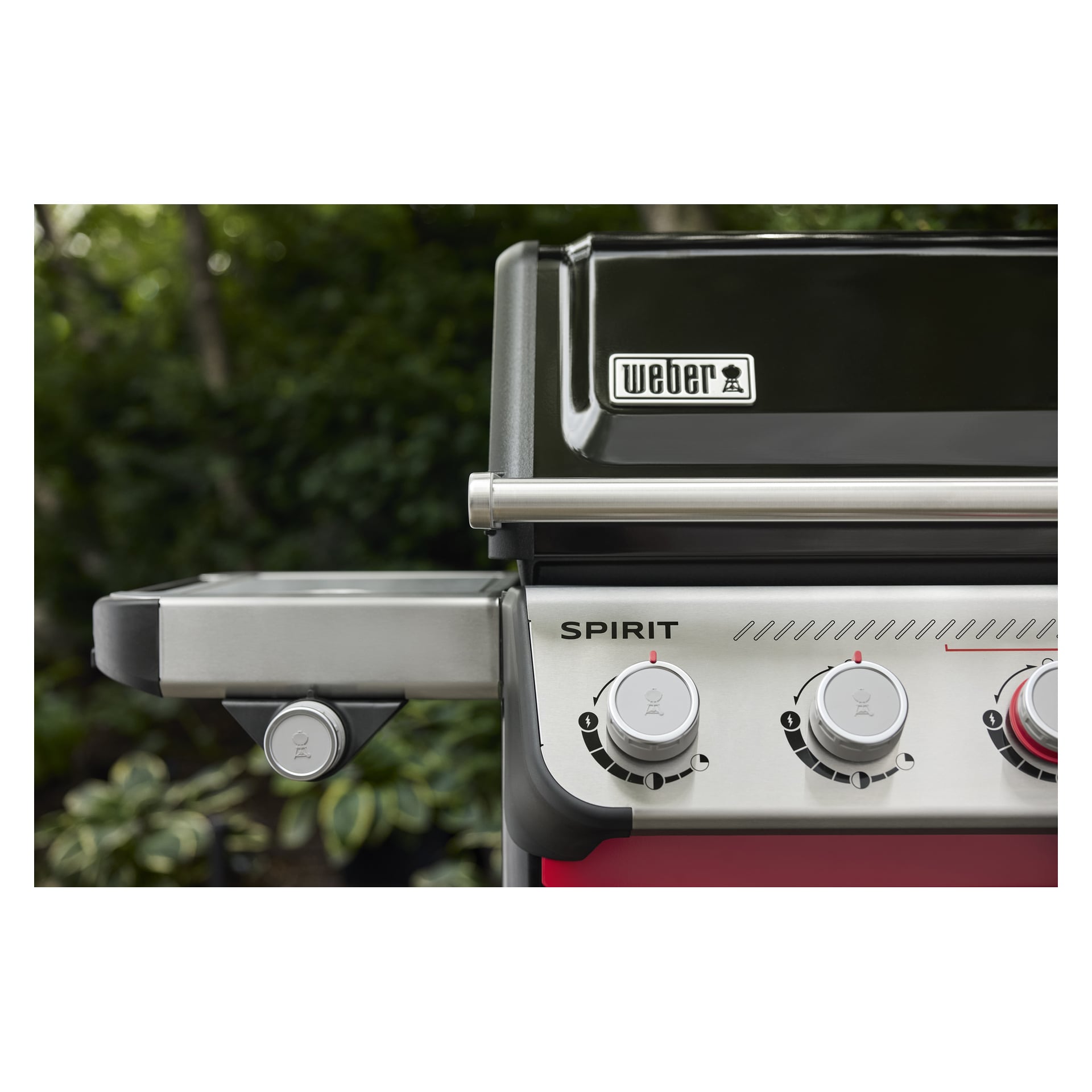 Detaljerad bild av Weber Spirits gasgrills kontrollrattar och sidohylla, bakgrunden mjukt suddig med grön växtlighet.