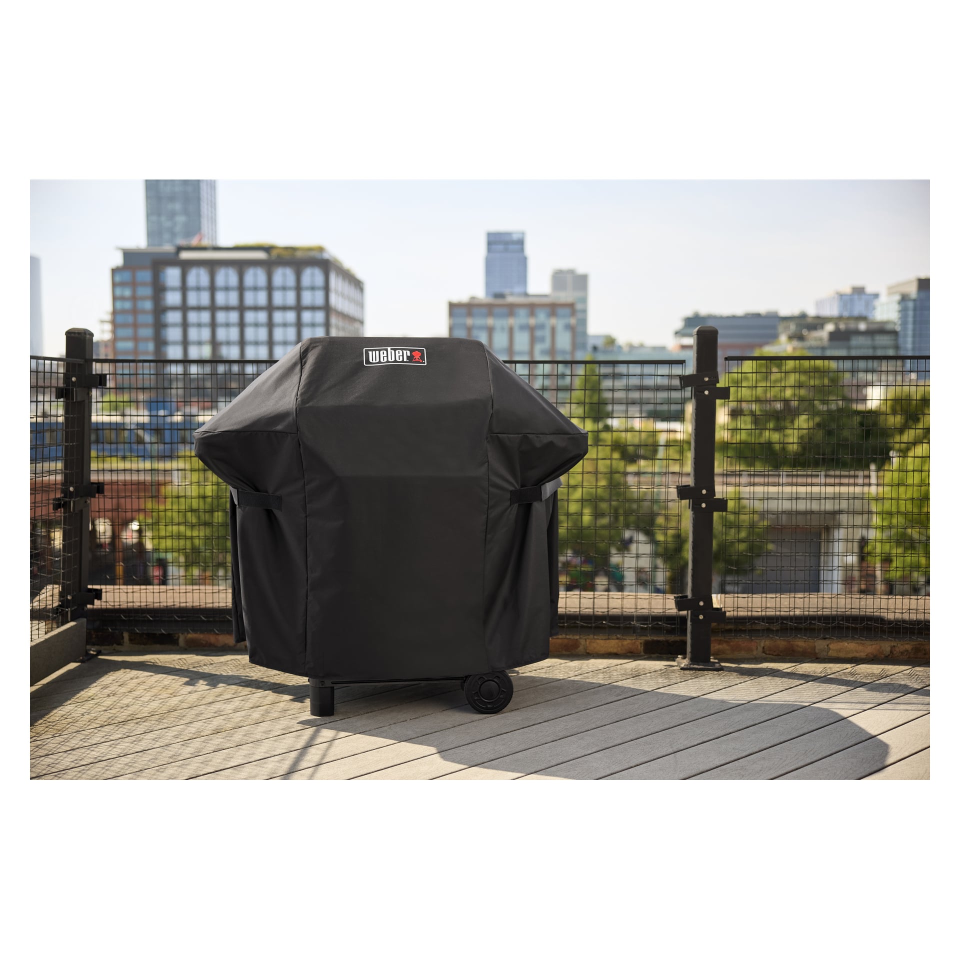 En svart Weber-grill med et tilpasset lokk står på en takterrasse nær et metallgjerde, med bybilde og grøntområder i bakgrunnen.