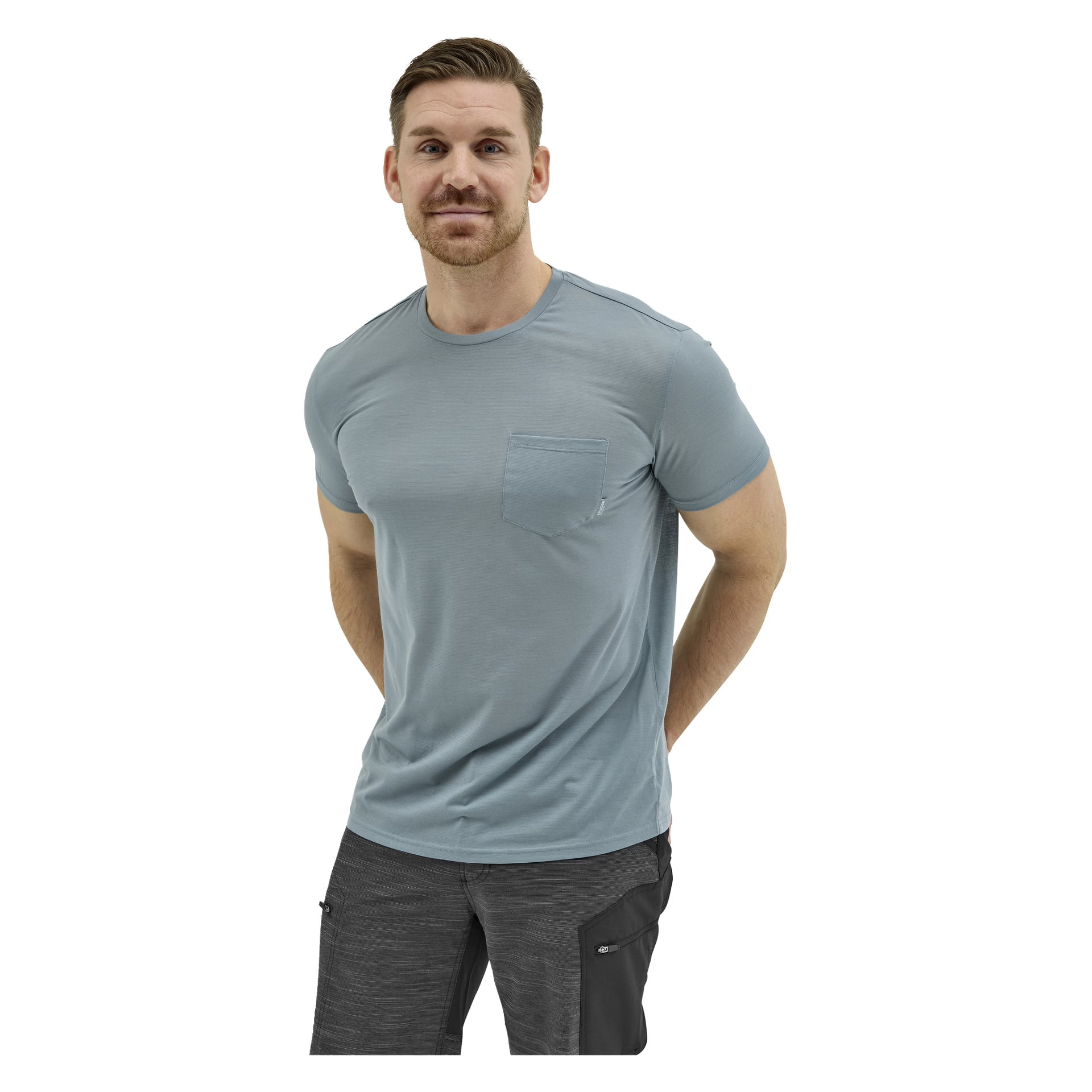 Ein Mann mit kurzen braunen Haaren, Bart und Schnurrbart trägt ein hellblaues Taschen-T-Shirt und dunkelgraue Hosen, die Hände hinter dem Rücken.