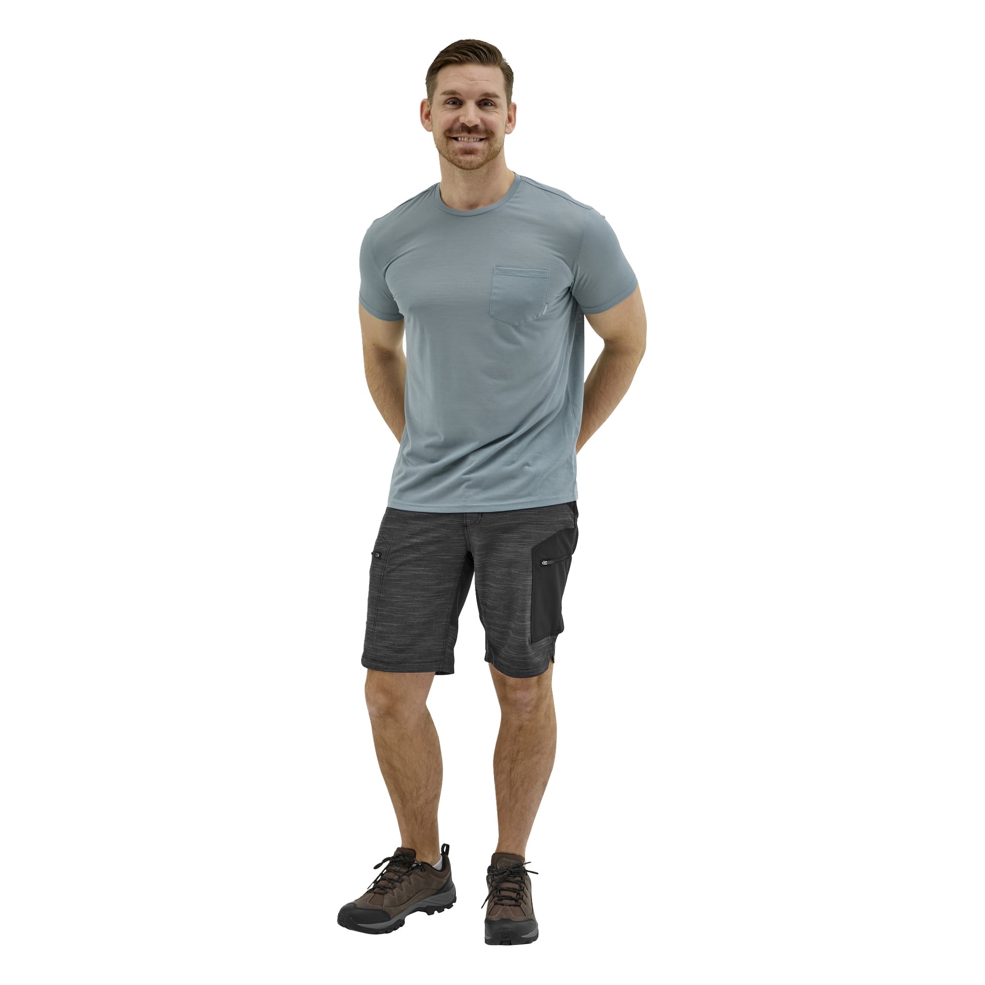 Ein Mann blickt nach vorne in einem hellblauen Kurzarmshirt, dunkelgrauen Cargoshorts und braunen Wanderschuhen, lächelt mit den Händen hinter dem Rücken.